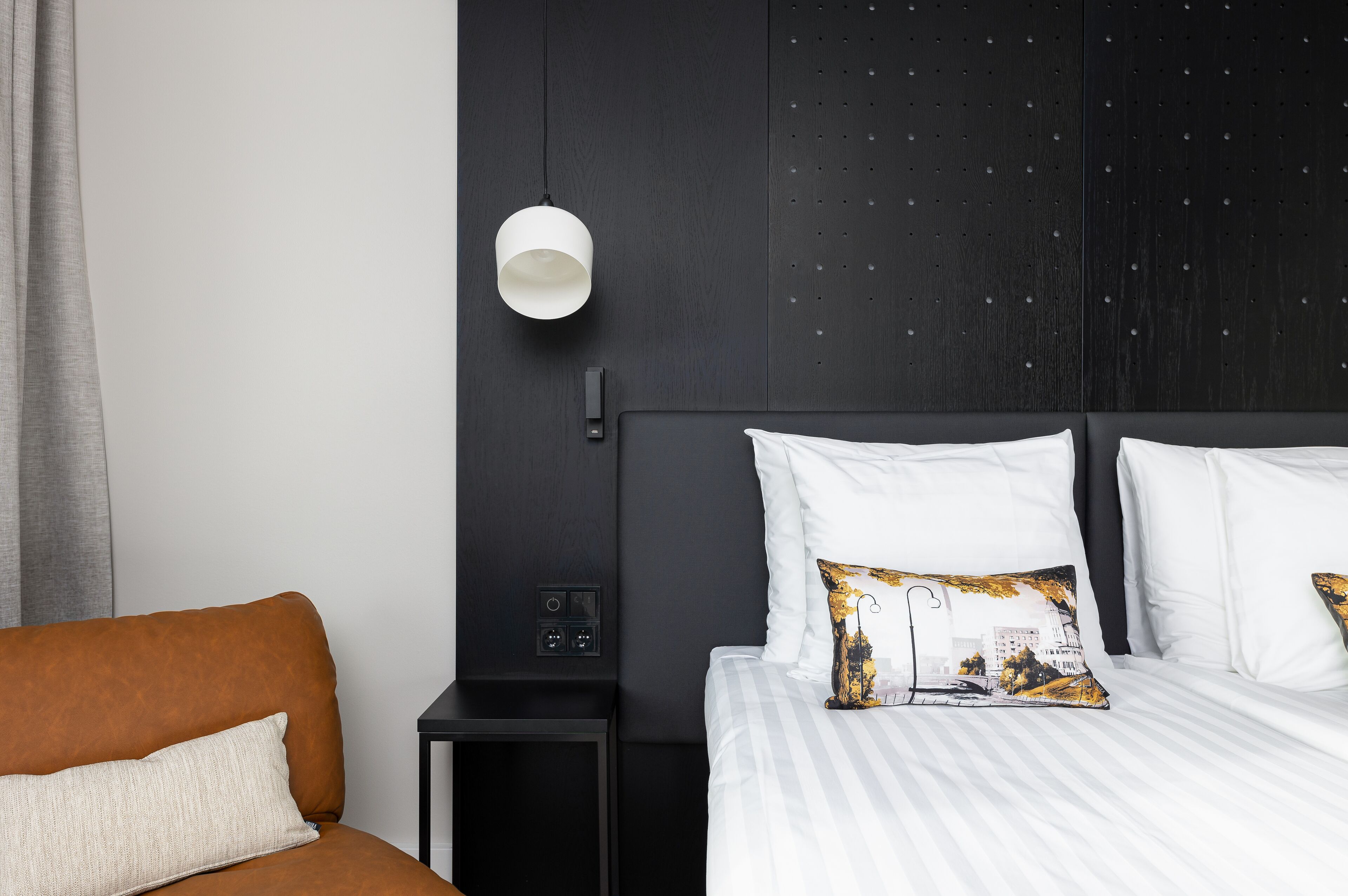 A modern hotel room with a large bed, a nightstand, a wall-mounted lamp, and a brown sofa.