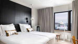 Quarto de hotel moderno com uma cama grande, uma mesa de trabalho e vista para a cidade através de uma janela ampla.