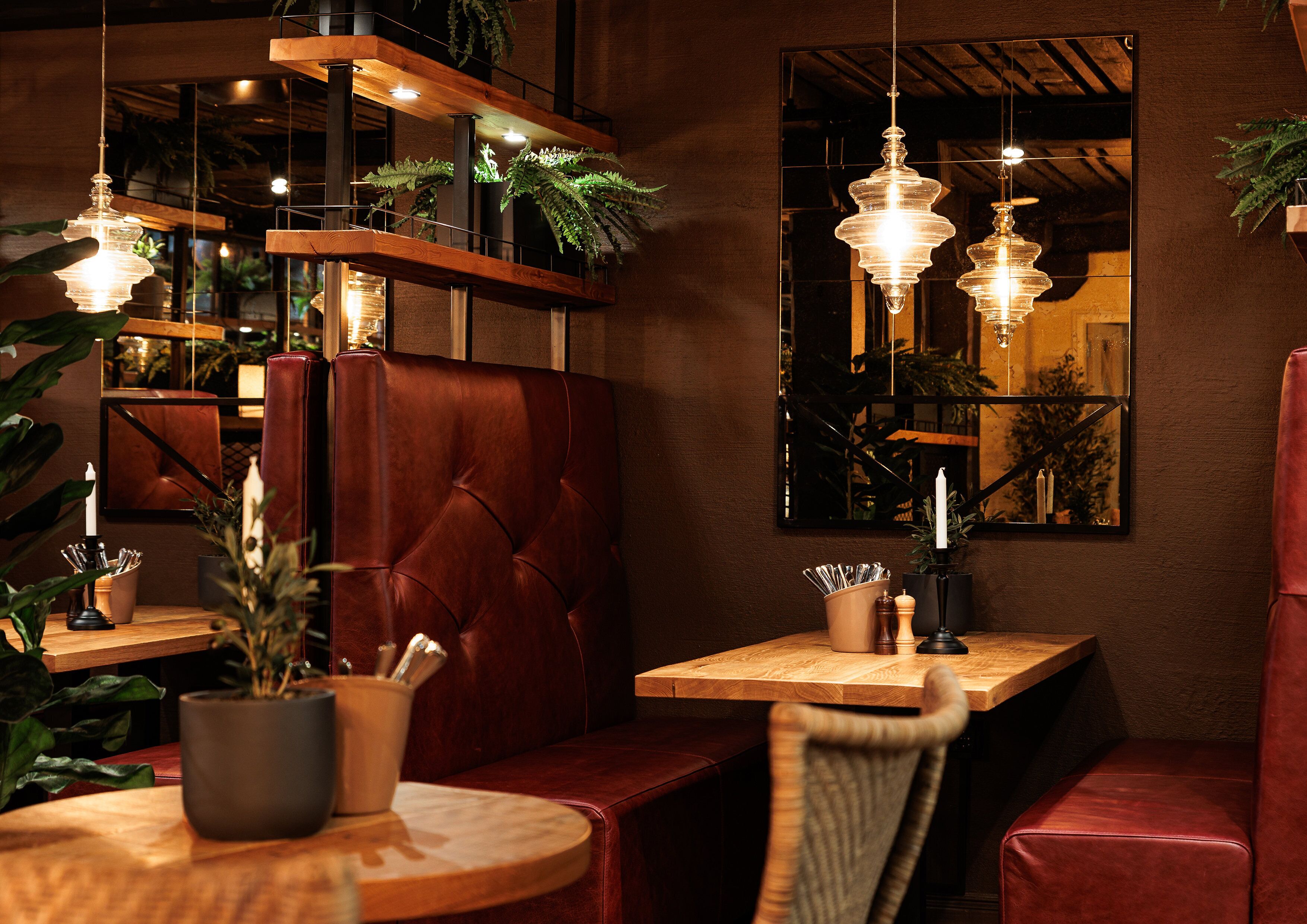 A cozy restaurant interior with leather booths, wooden tables, hanging pendant lights, and potted plants.