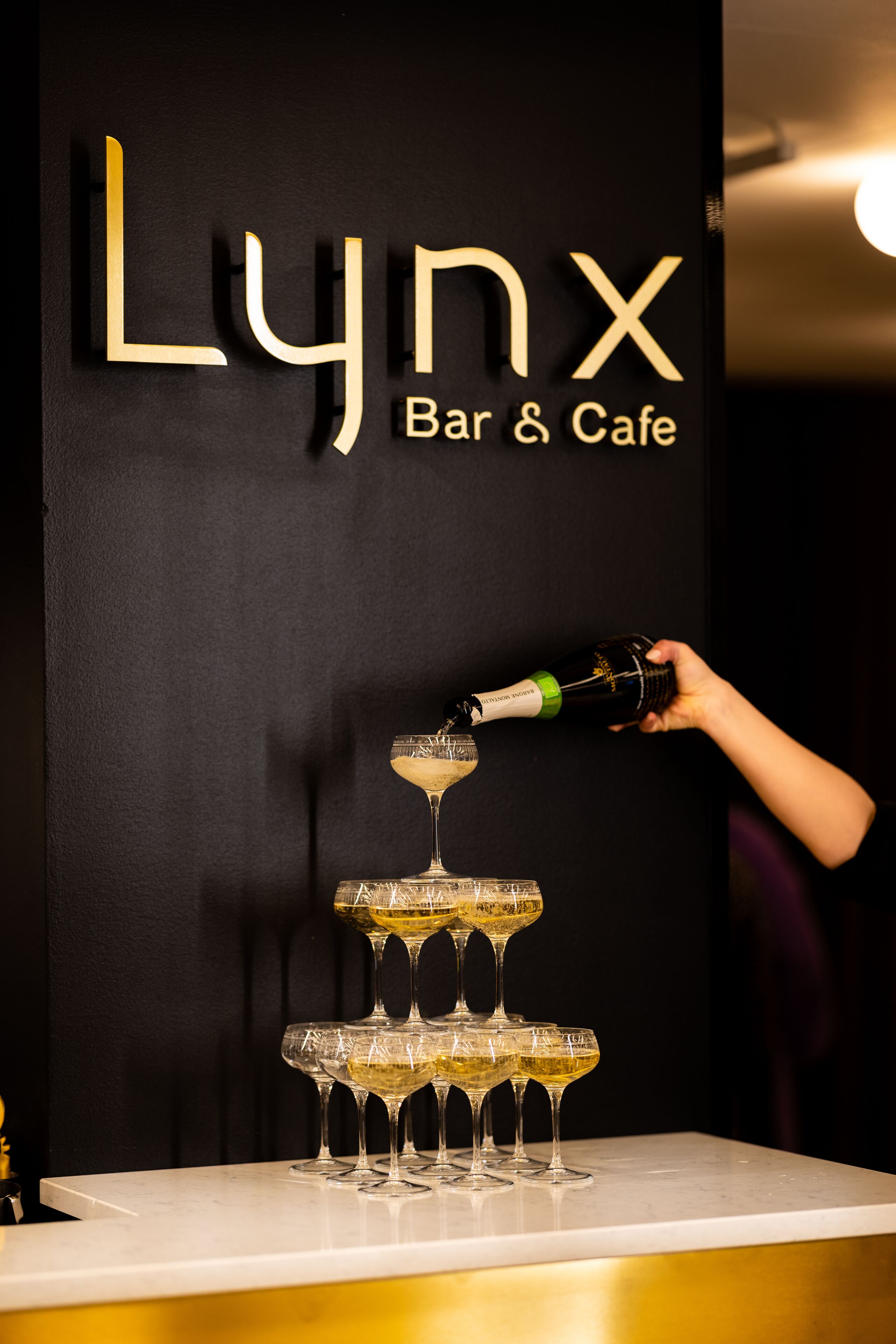 A bar setup with a sign reading "LYNX Bar & Cafe" and a hand pouring a drink into a glass.