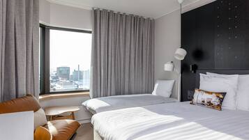 Une chambre d’hôtel moderne avec deux lits, une petite table et une vue sur le paysage urbain grâce à de grandes fenêtres.