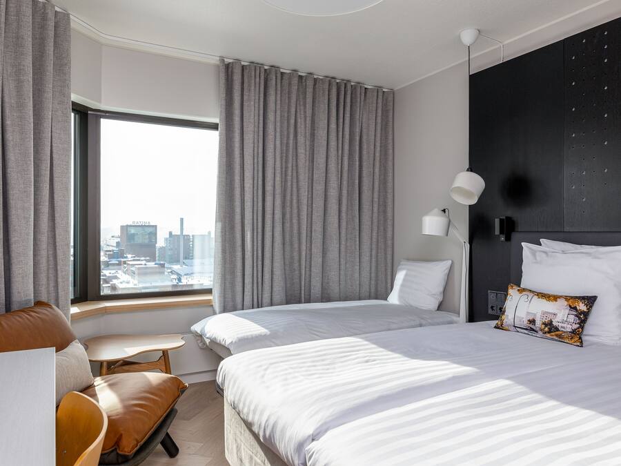 Une chambre d’hôtel moderne avec deux lits, une petite table et une vue sur le paysage urbain grâce à de grandes fenêtres.