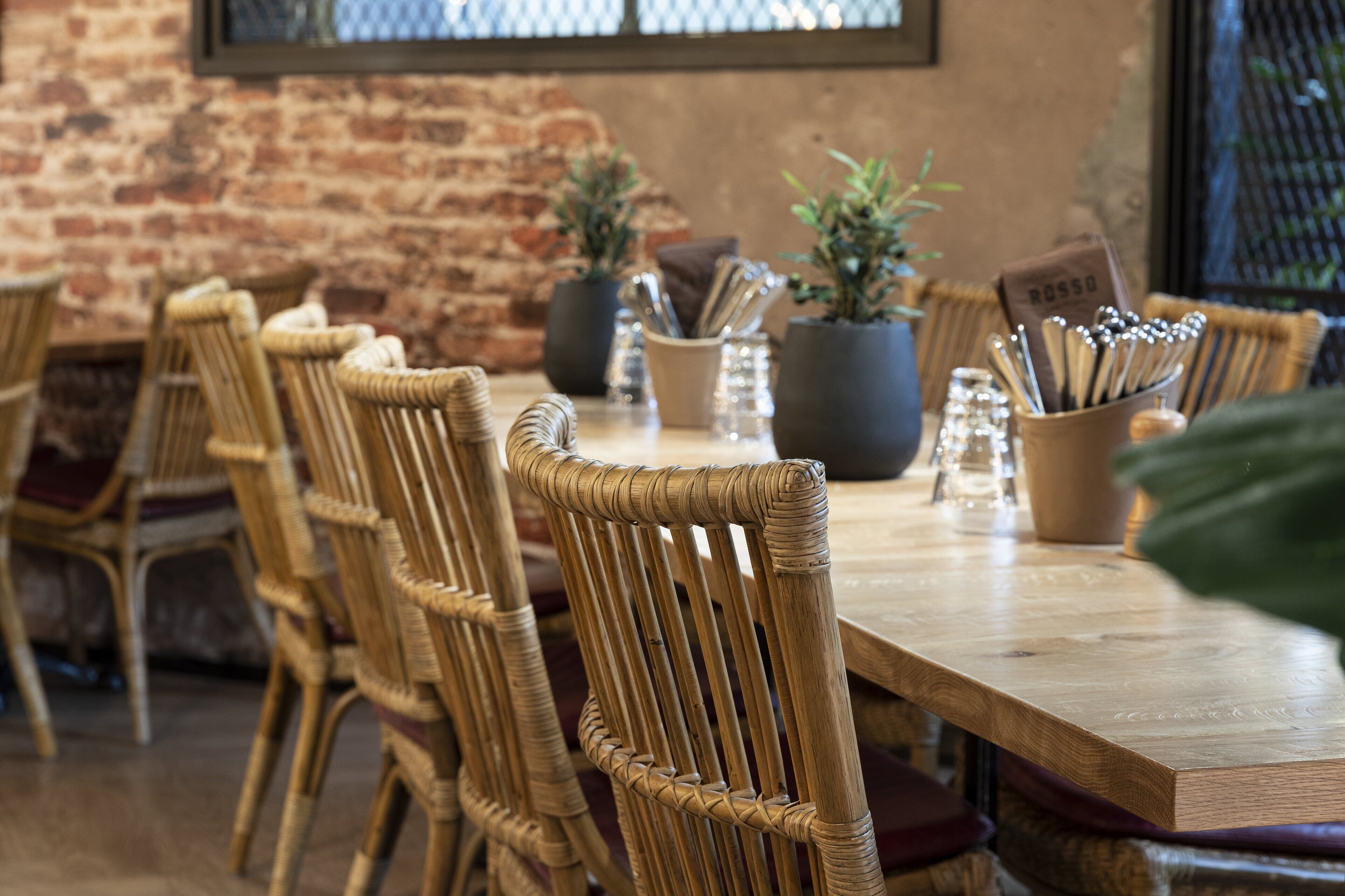Une table à manger en bois, entourée de chaises en osier, agrémentée de plantes en pot et d’un ensemble de couverts posé sur la table.