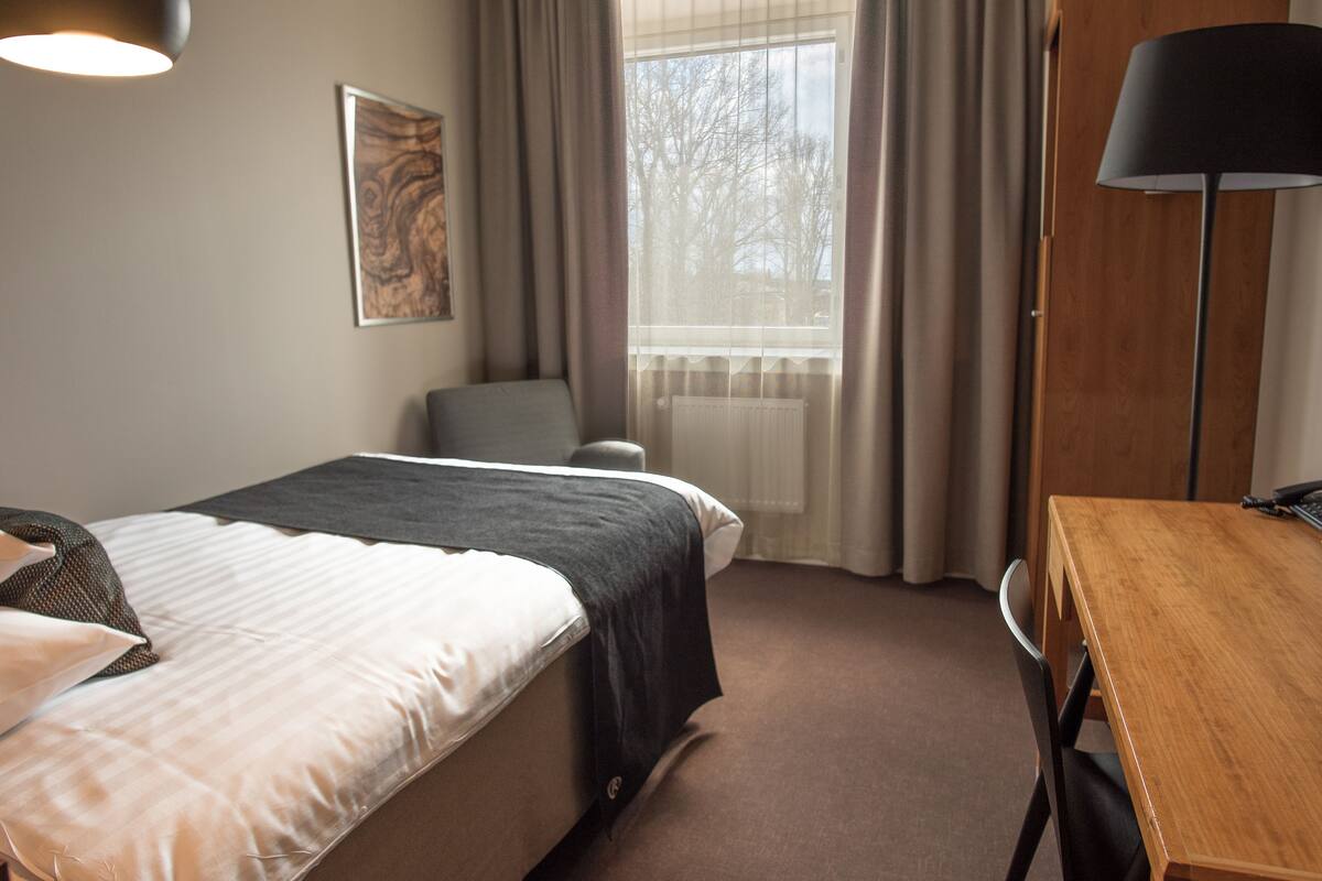 A hotel room with a bed, a desk, a chair, a lamp, and a window with curtains.
