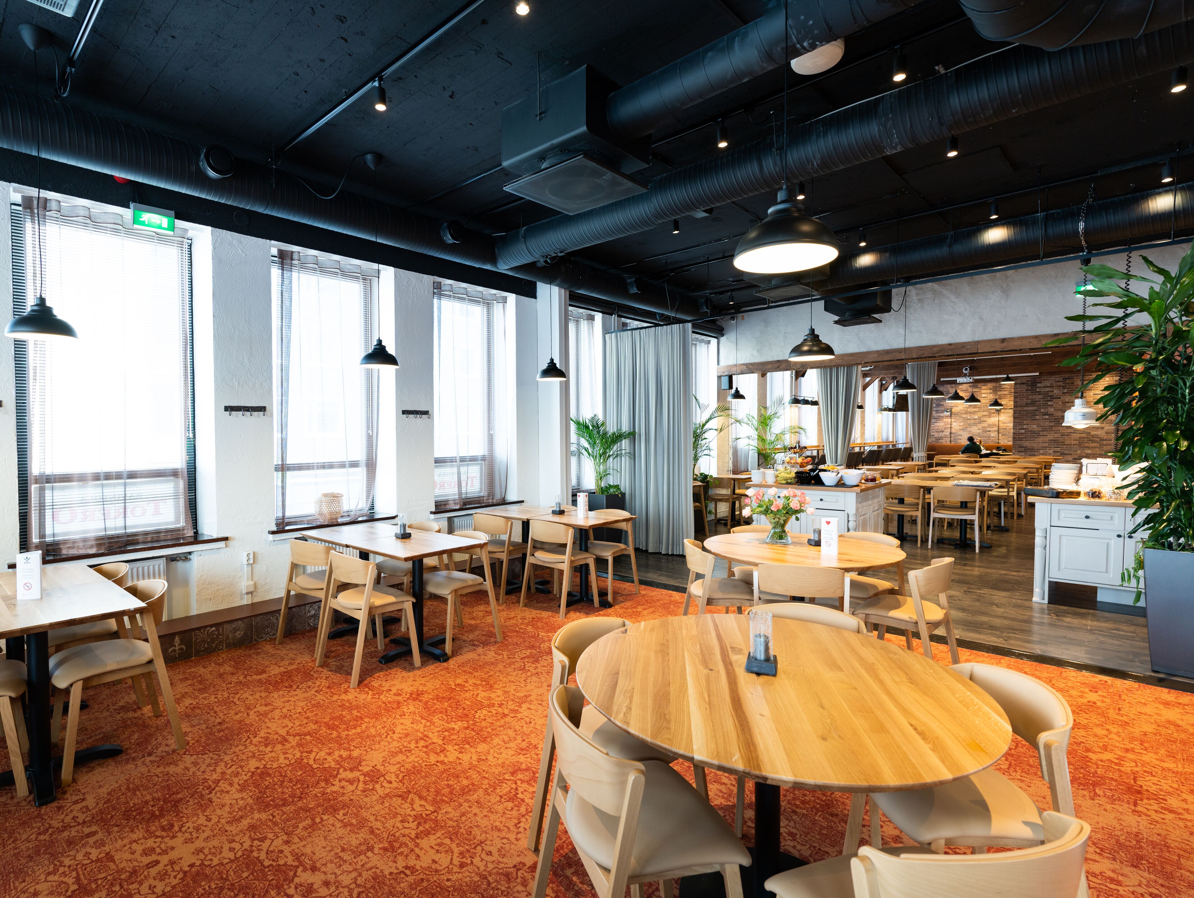 A modern dining area with multiple tables and chairs, large windows, and a kitchen in the background.