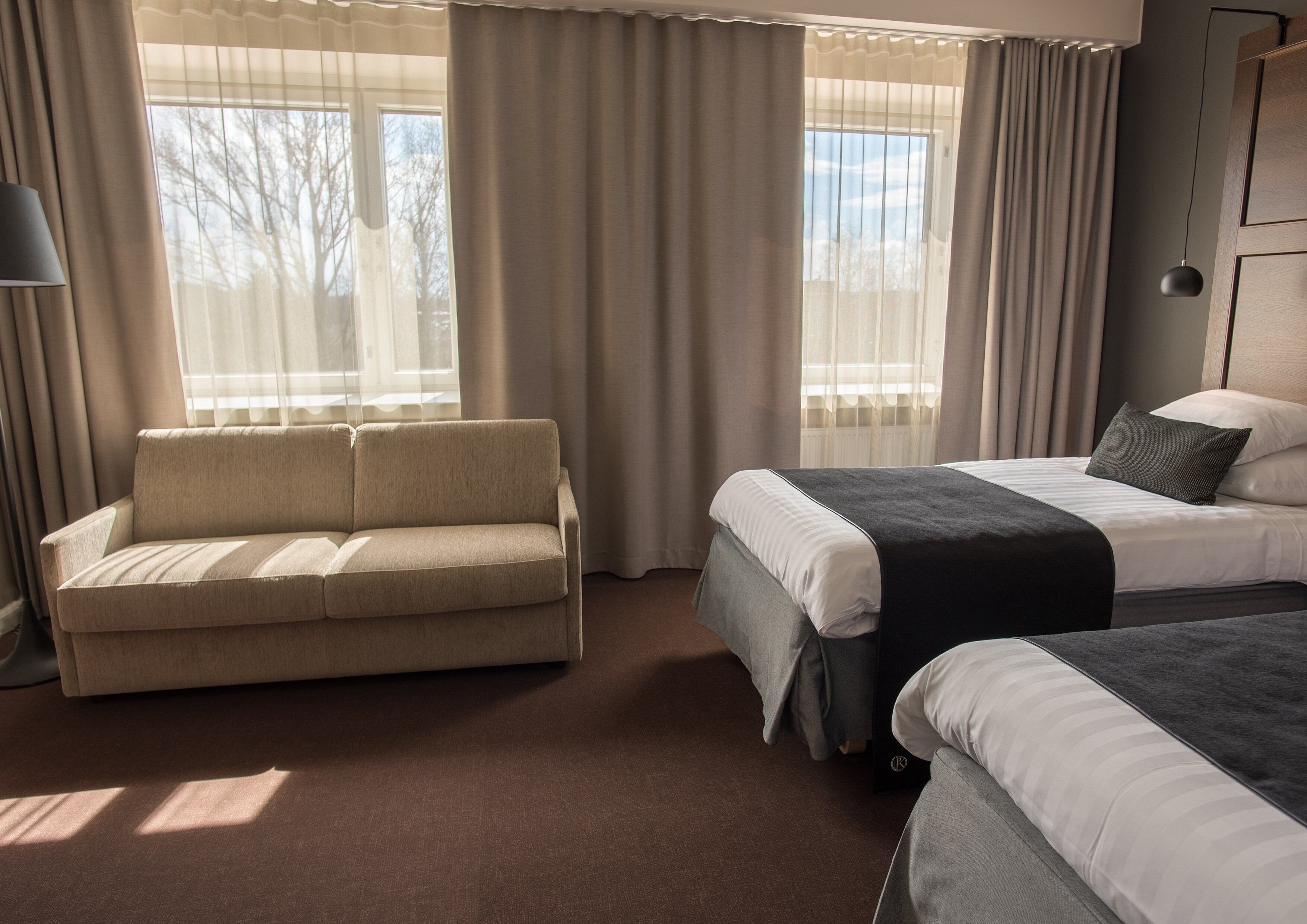 A hotel room with a sofa, two beds, and large windows with curtains.