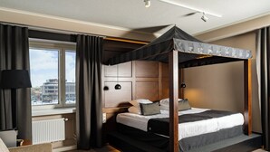 A modern hotel room with a four-poster bed, a view of the city through large windows, a cozy seating area, and a well-lit ambiance. - Original Sokos Hotel Vaakuna Joensuu (Joensuu)