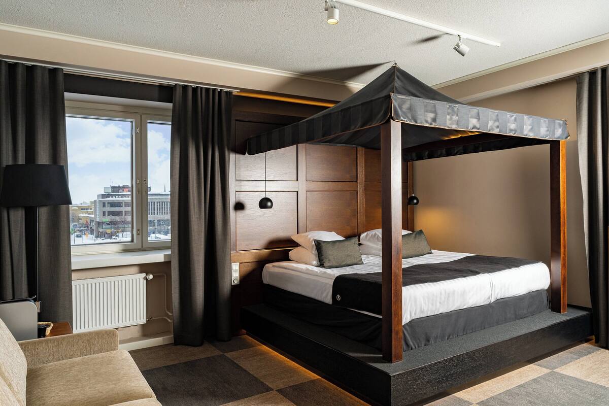 A modern hotel room with a four-poster bed, a view of the city through large windows, a cozy seating area, and a well-lit ambiance.