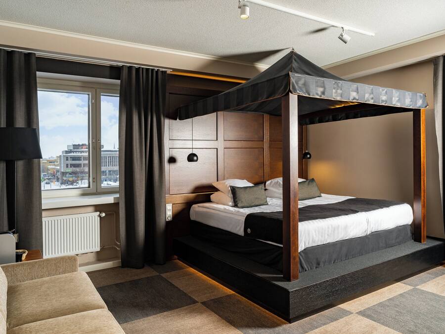Una habitación de hotel moderna con una cama con dosel, vistas de la ciudad a través de amplias ventanas, un rincón de descanso acogedor y un ambiente luminoso.