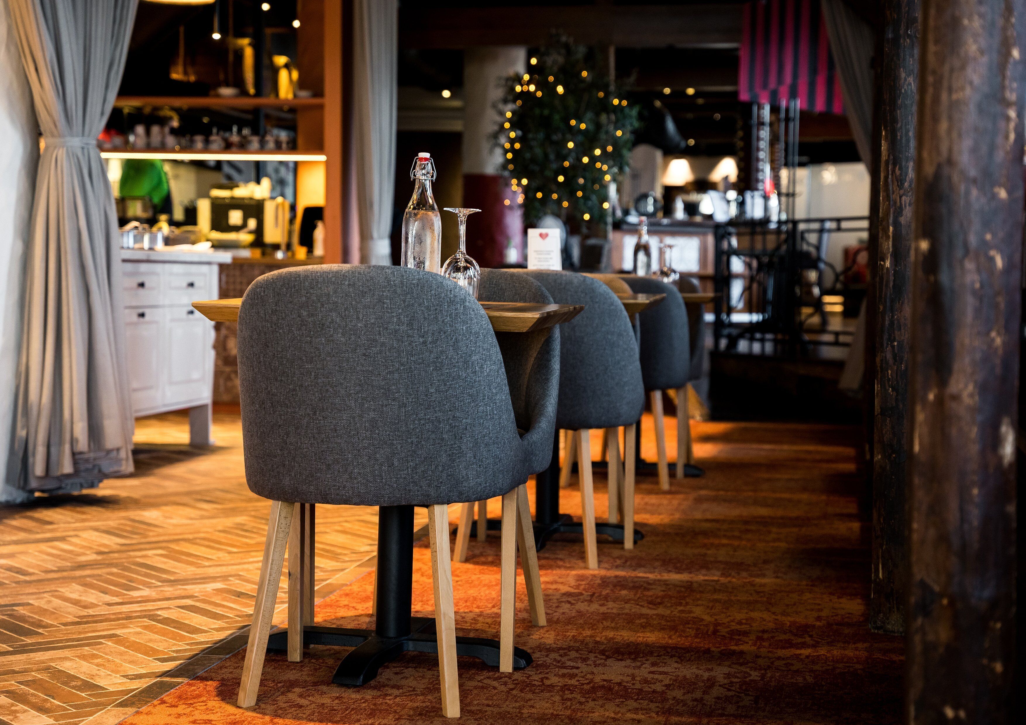 Interior de restaurante con mesas de madera y sillas tapizadas en gris, una botella de vino y un árbol de Navidad al fondo.
