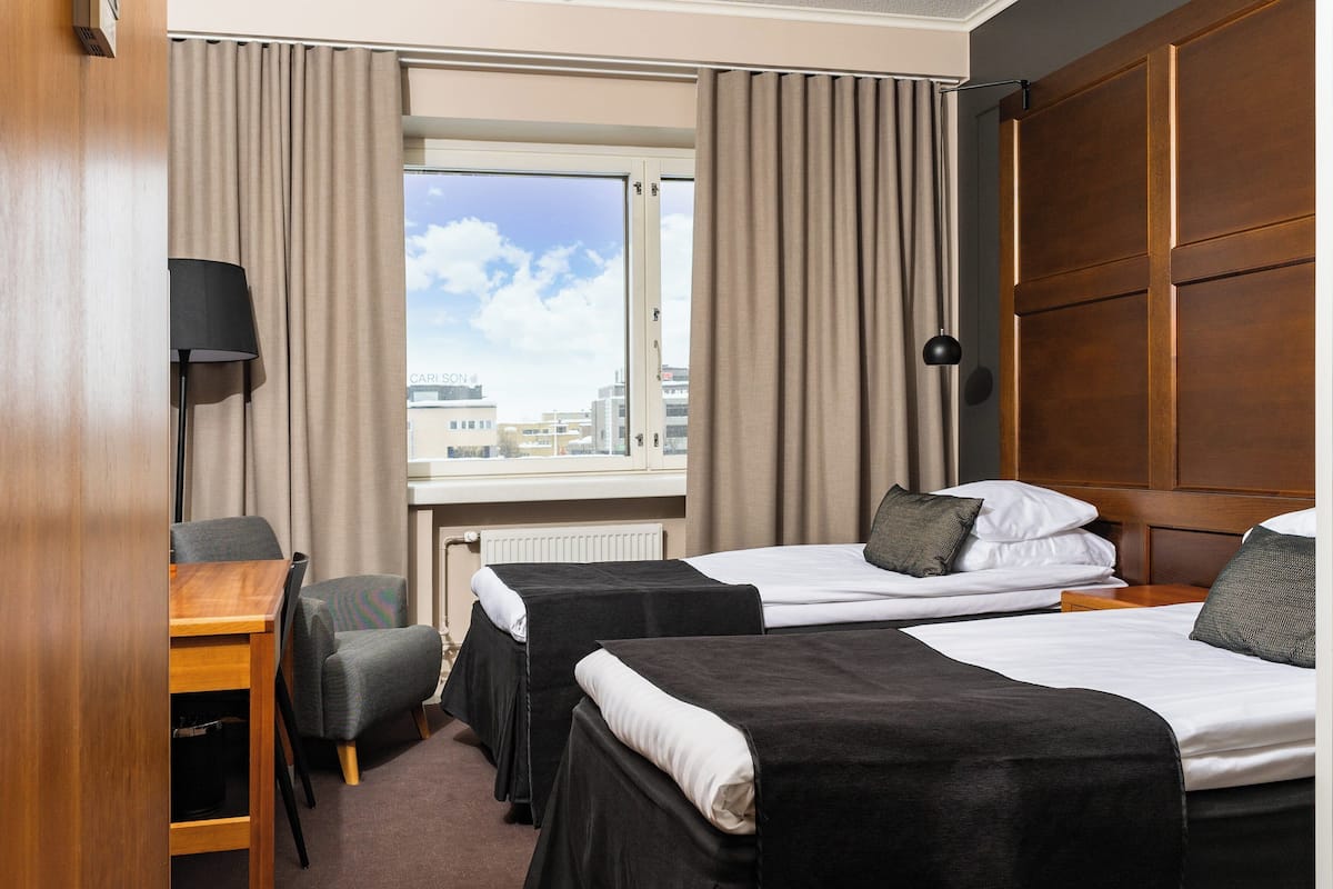 A hotel room with two beds, a desk, a chair, a lamp, and a window with curtains.