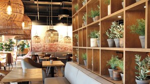 Un café acogedor con mesas y bancos de madera, lámparas de mimbre colgantes y una pared de ladrillo con plantas en macetas.