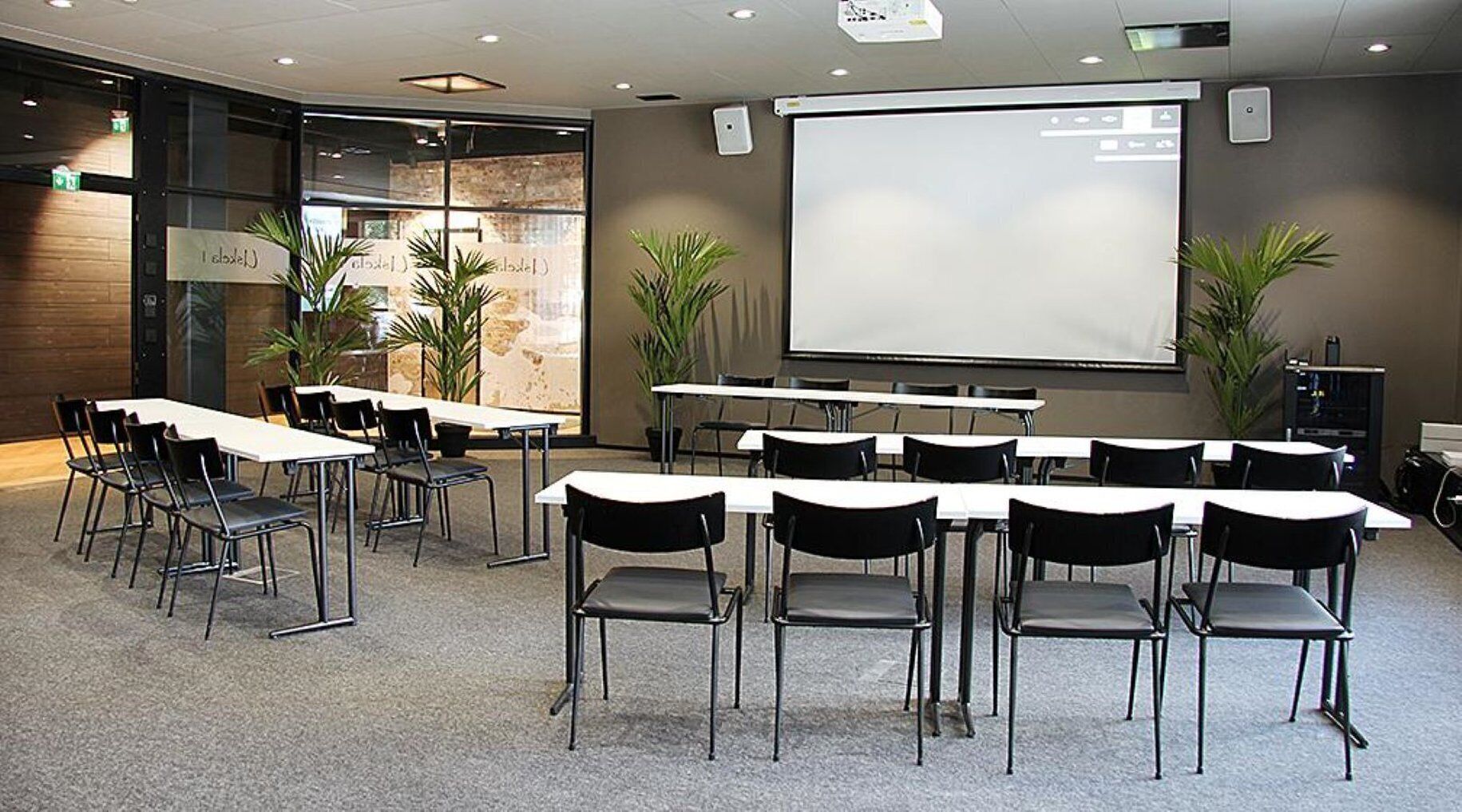 Een vergaderruimte met tafels, stoelen, een scherm voor een projecter en potplanten.