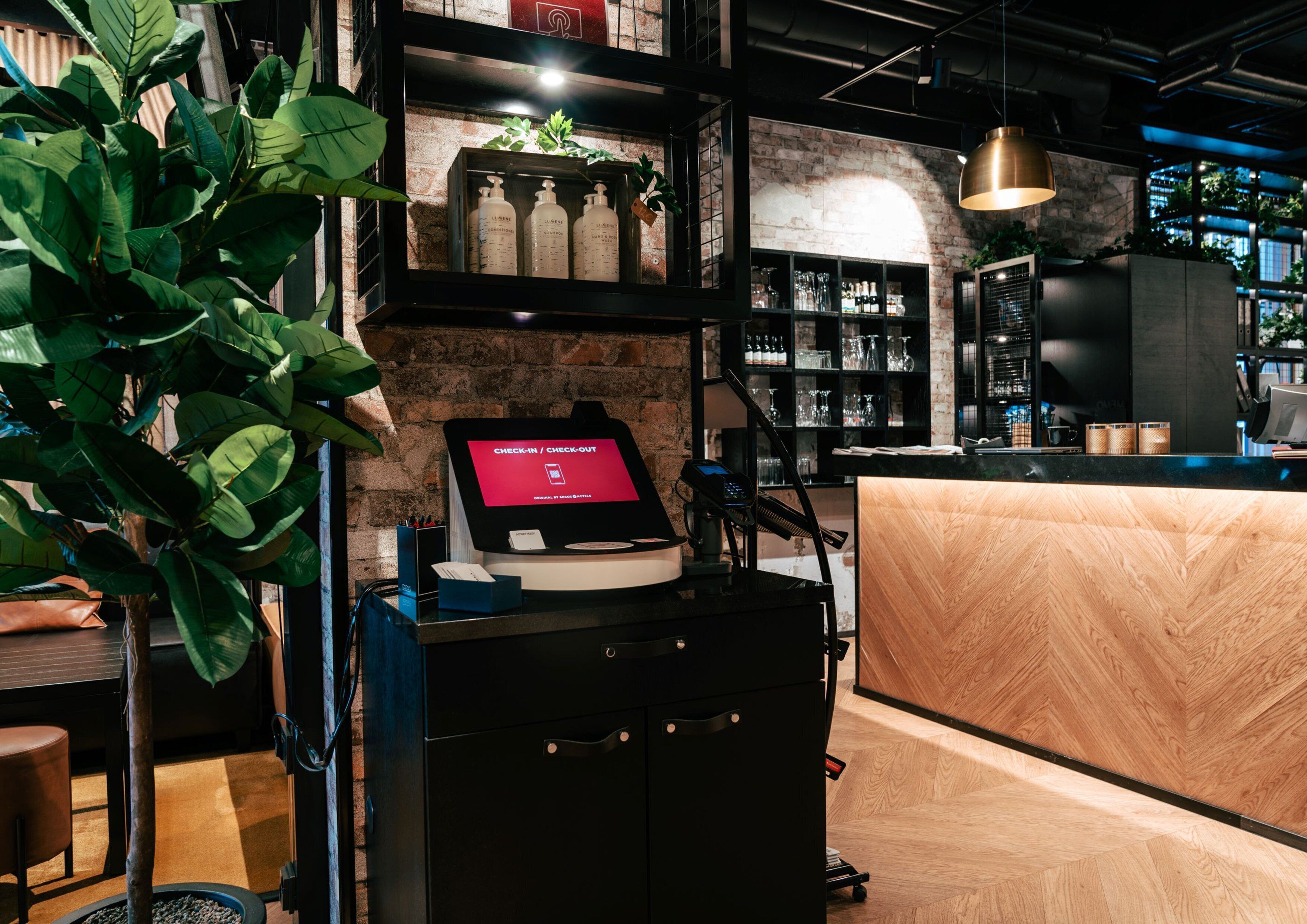 A check-in/check-out kiosk, a wooden reception desk, and a brick wall with shelves in a modern interior.