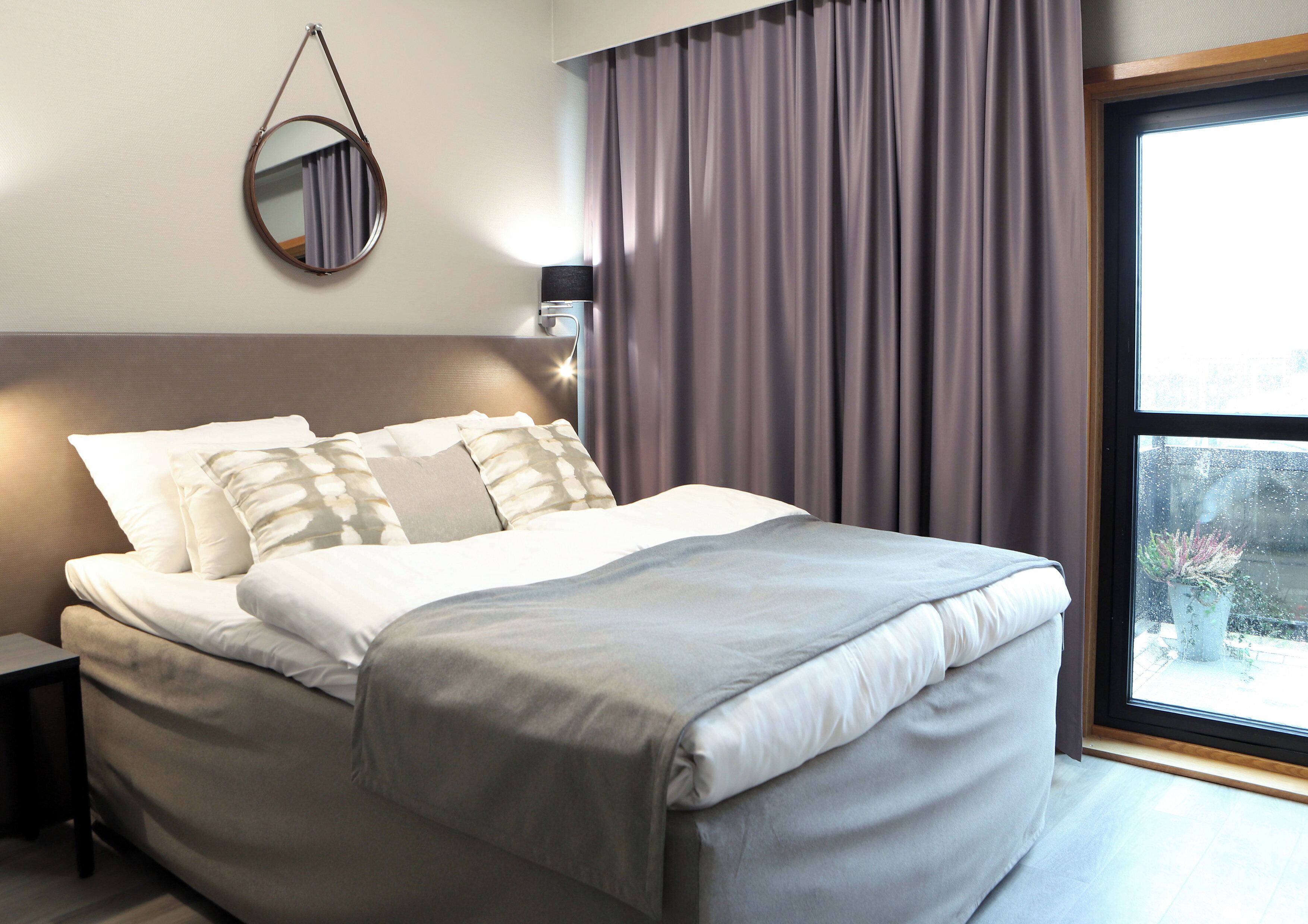 A neatly made bed with a grey duvet, a bedside lamp, and a round mirror on the wall.