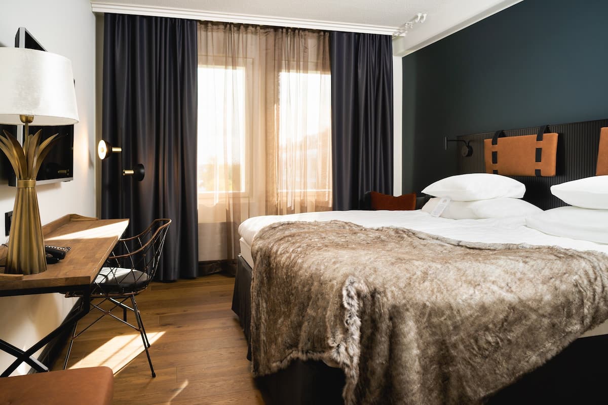 Une chambre d’hôtel avec un grand lit, un bureau avec une lampe, une chaise et une fenêtre avec des rideaux.