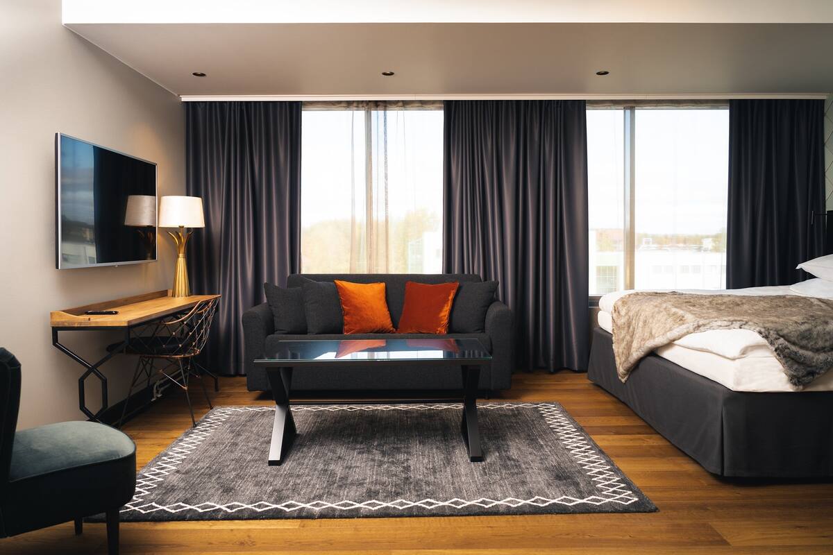 Une chambre d’hôtel moderne, dotée d’un canapé, d’un bureau avec une lampe, d’un lit avec une couverture et de grandes fenêtres avec des rideaux.
