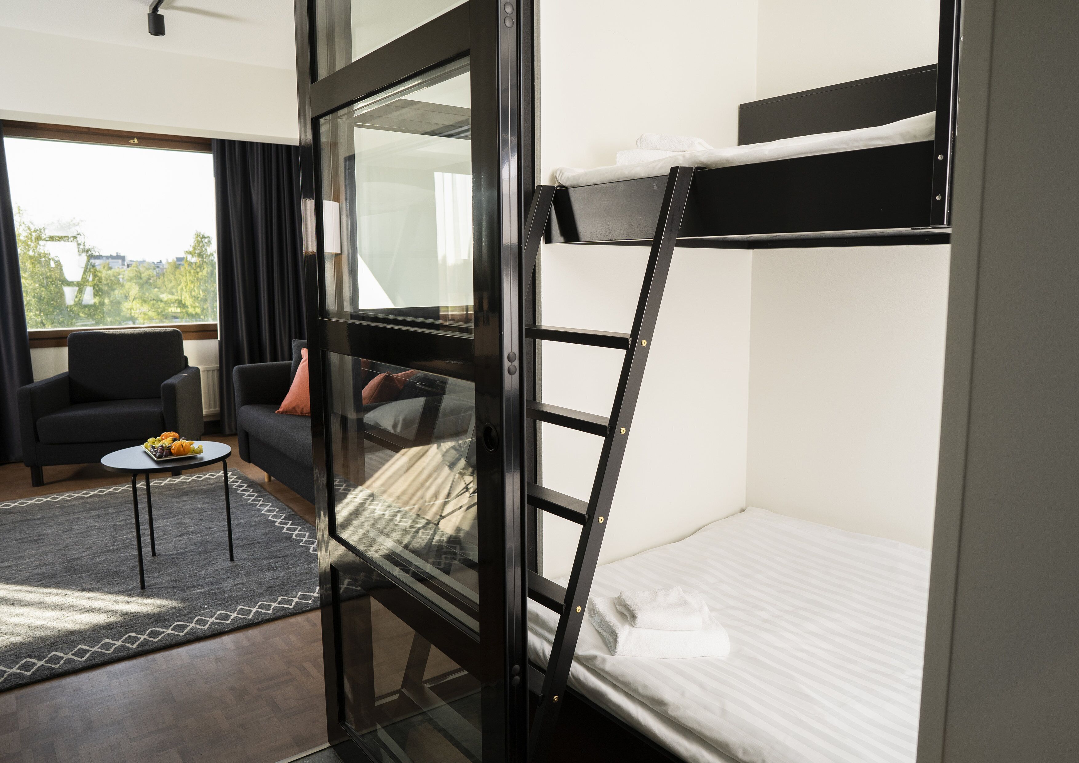 A modern room with a bunk bed, a sofa, a small table with fruit, and a view of greenery outside.