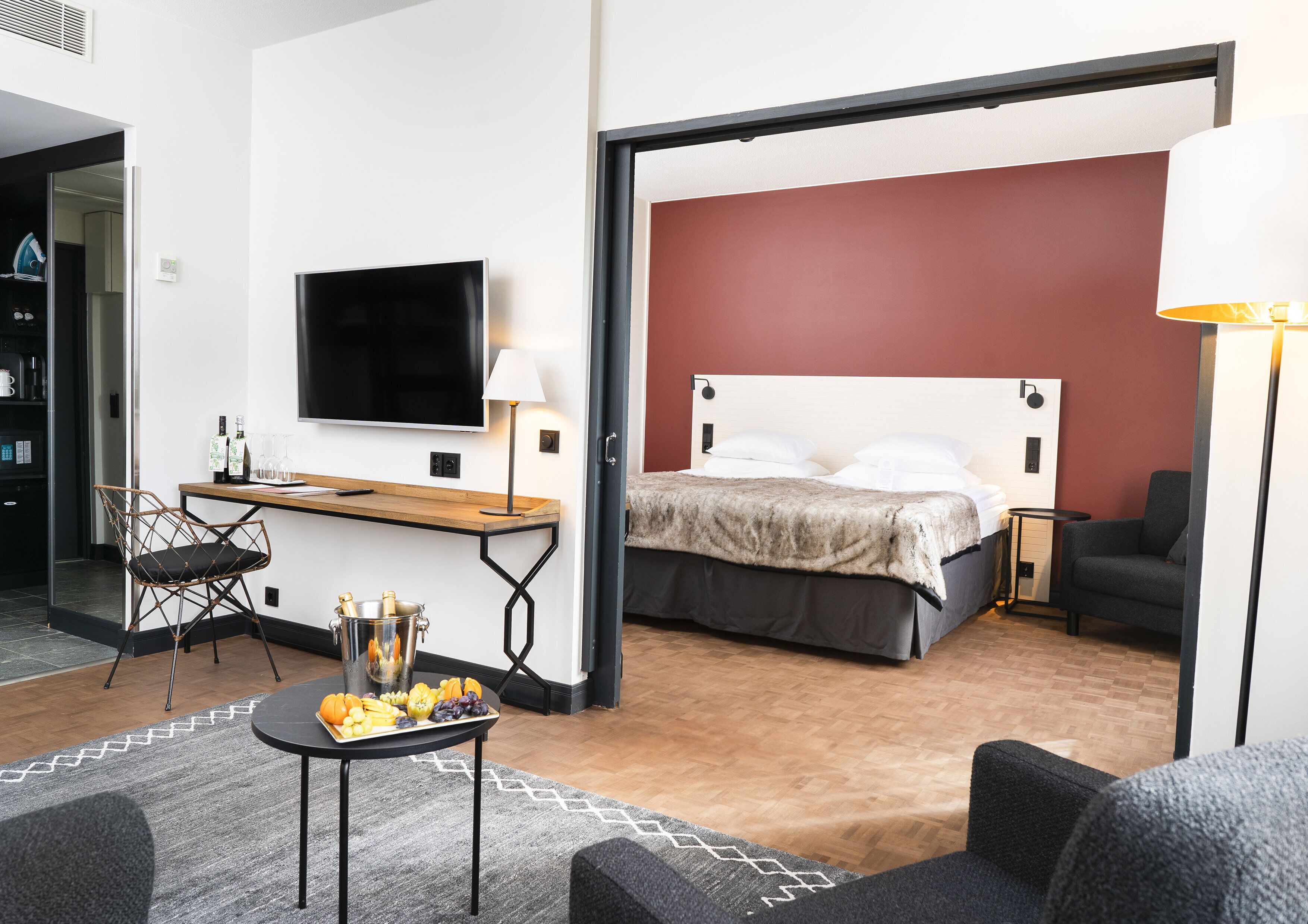 A hotel room with a bed, a desk with a TV, a chair, a table with fruit, and a lamp.