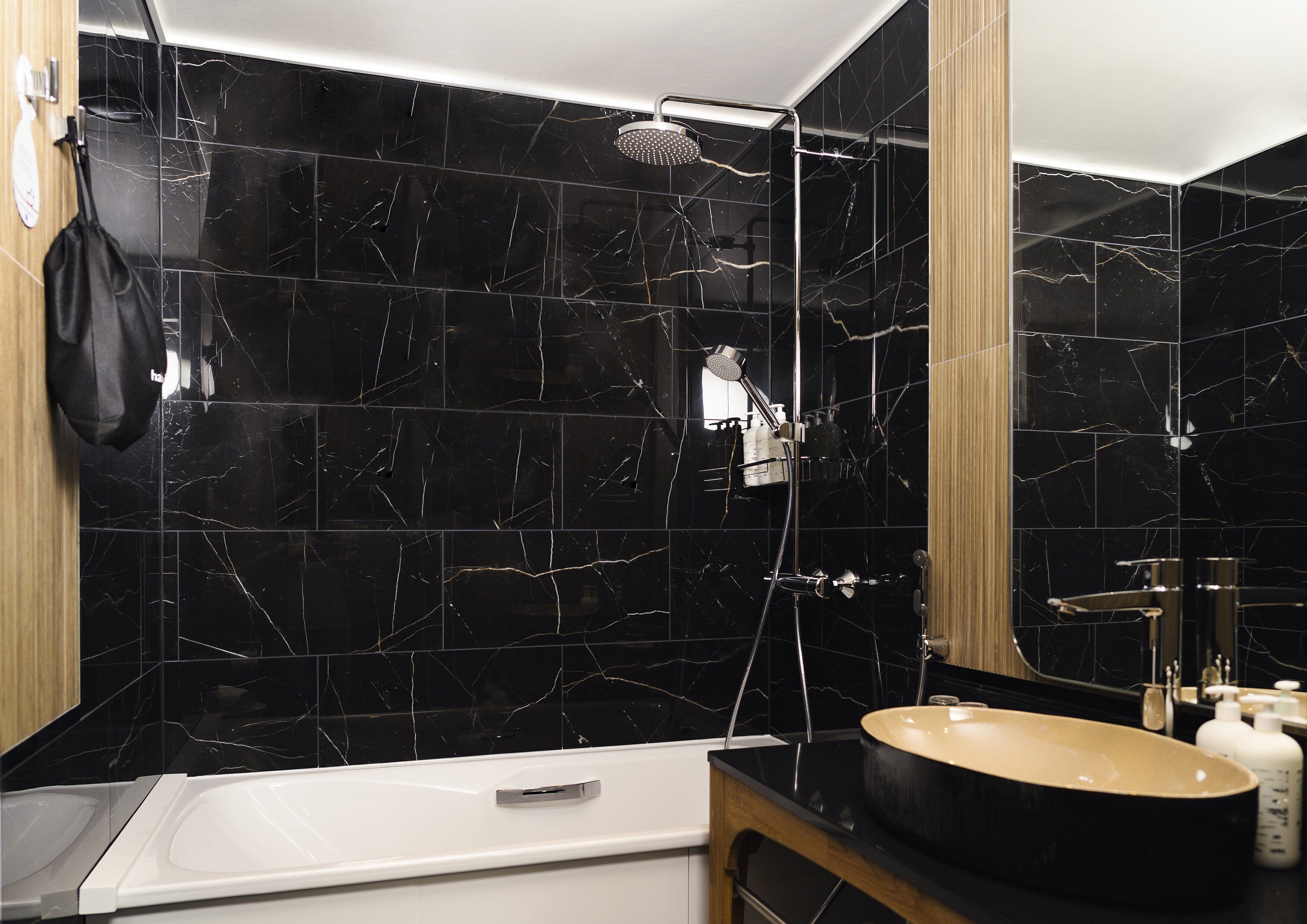 A modern bathroom with black marble tiles, a white bathtub, and a black sink with a beige basin.