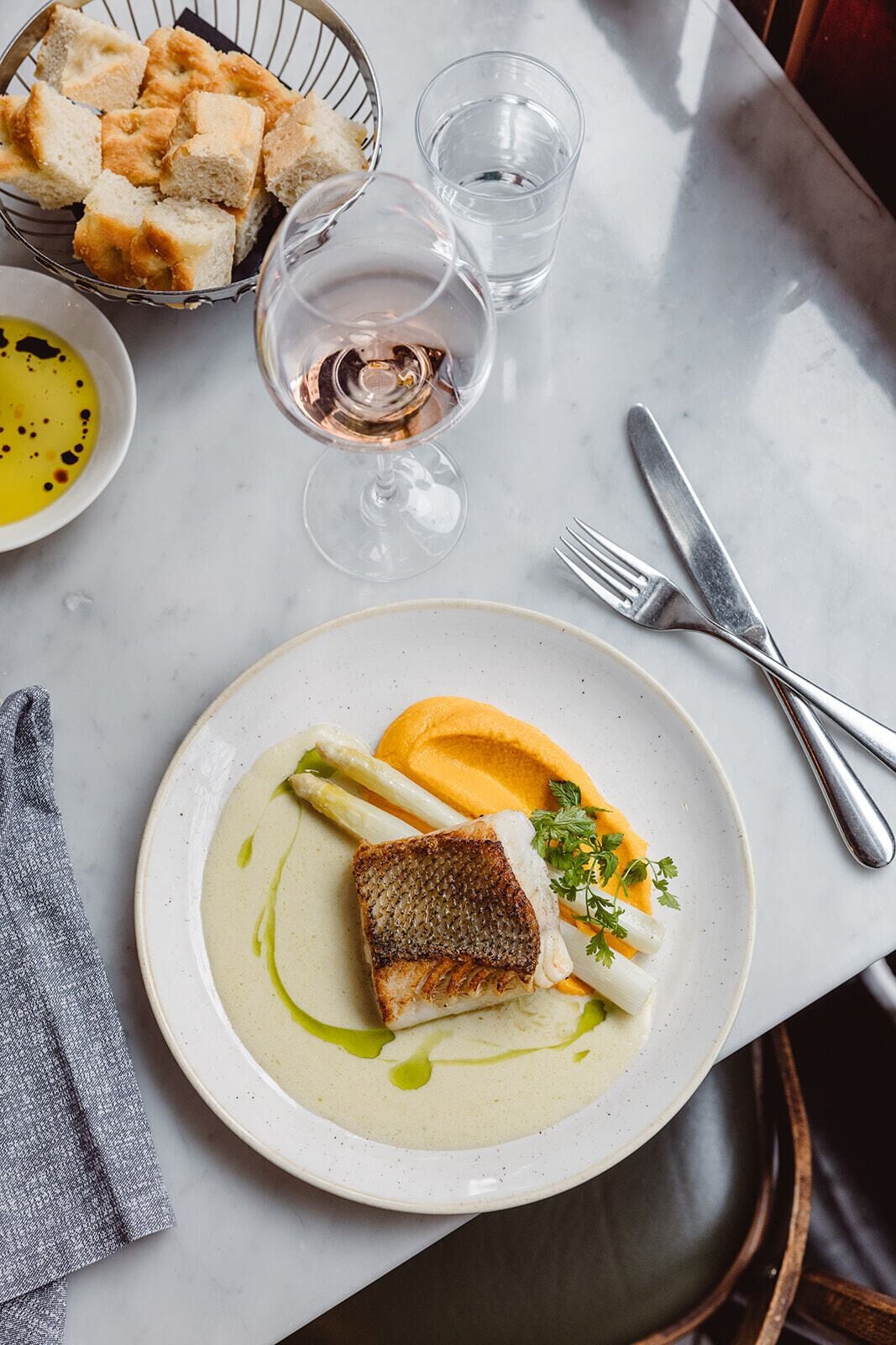 A dining table with a plate of fish, asparagus, and sauce, a glass of wine, and a basket of bread.