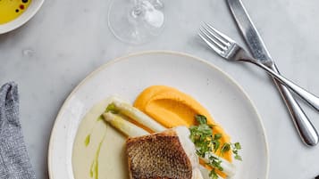 A dining table with a plate of fish, asparagus, and sauce, a glass of wine, and a basket of bread.