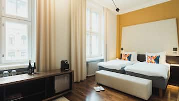 Habitación de hotel con piso de madera, cama con ropa blanca y almohadas coloridas, un banco, una mesita con botellas y vista a un edificio por la ventana.