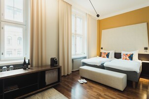 Habitación de hotel con piso de madera, cama con ropa blanca y almohadas coloridas, un banco, una mesita con botellas y vista a un edificio por la ventana.