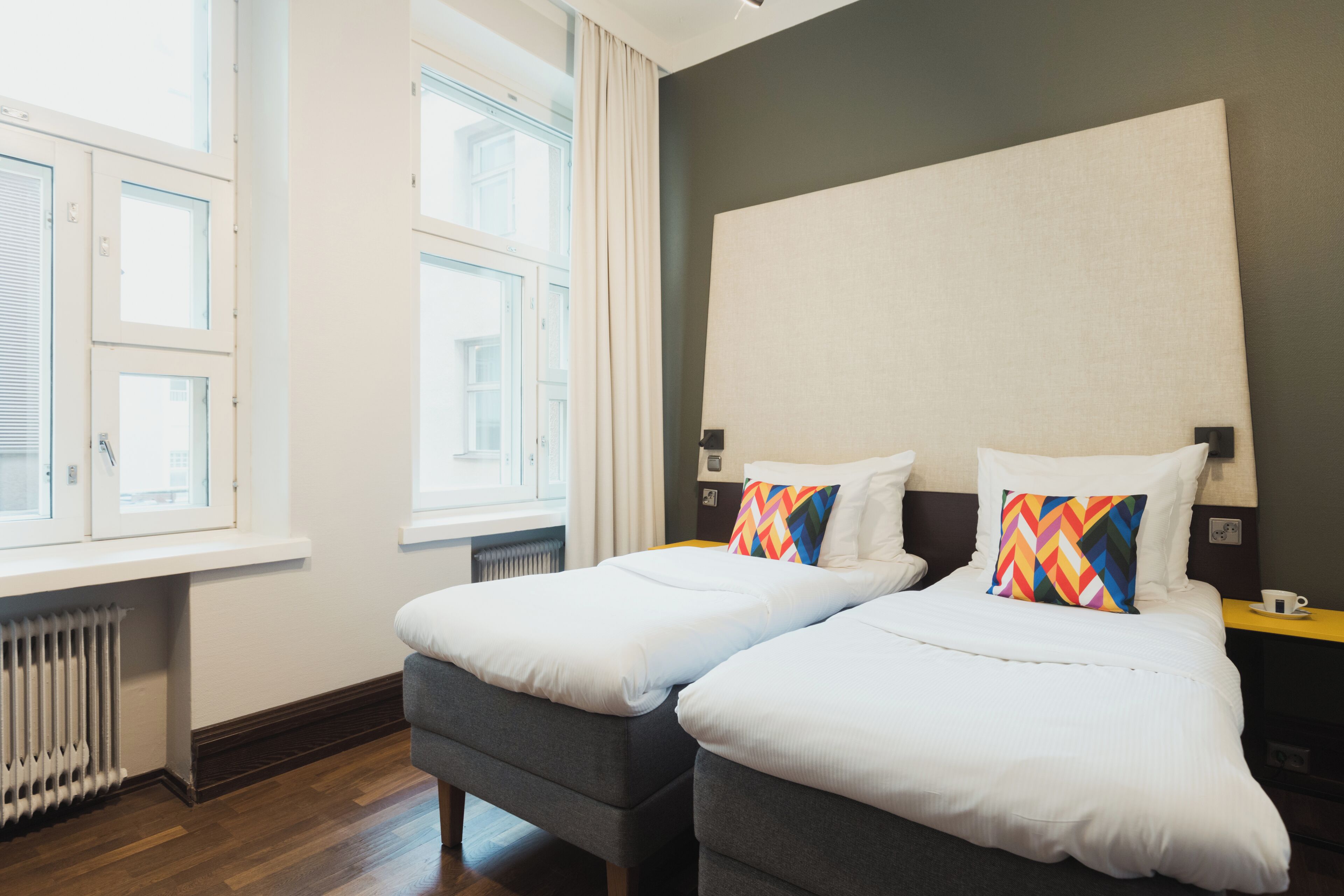 A hotel room with two beds, a radiator, a window with curtains, and a view of the outside.