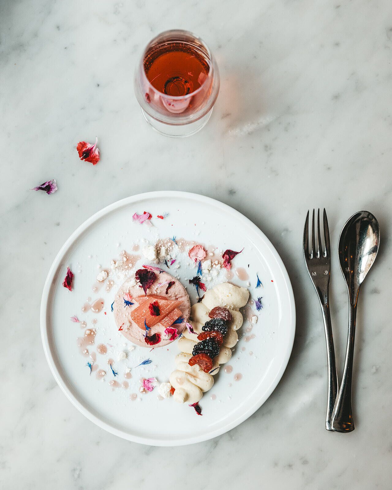 A white plate with a dessert, a glass of red liquid, and a fork and spoon on a marble surface.