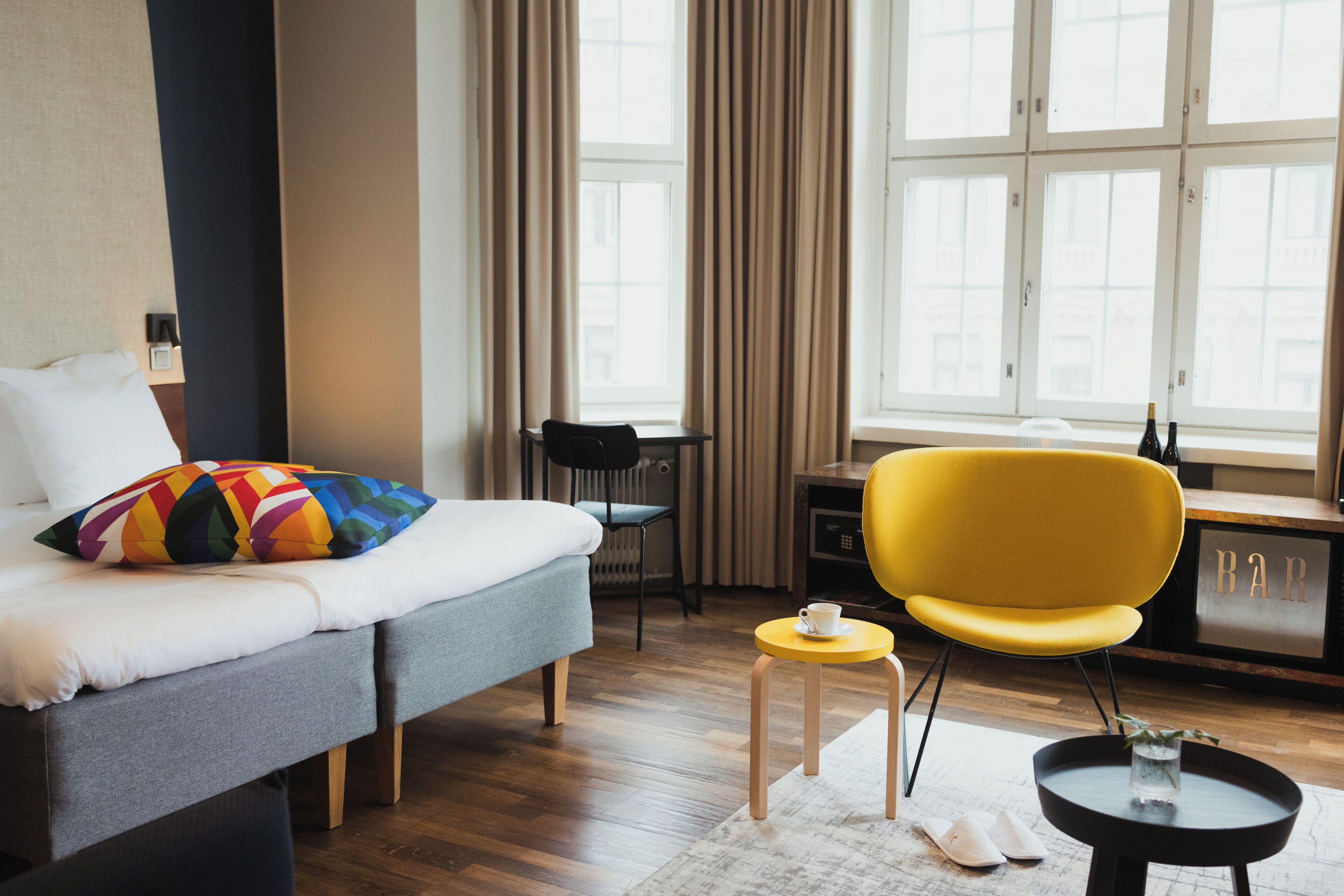 Une chambre d’hôtel avec un lit, une petite table, une chaise jaune, un bureau et une fenêtre avec des rideaux.