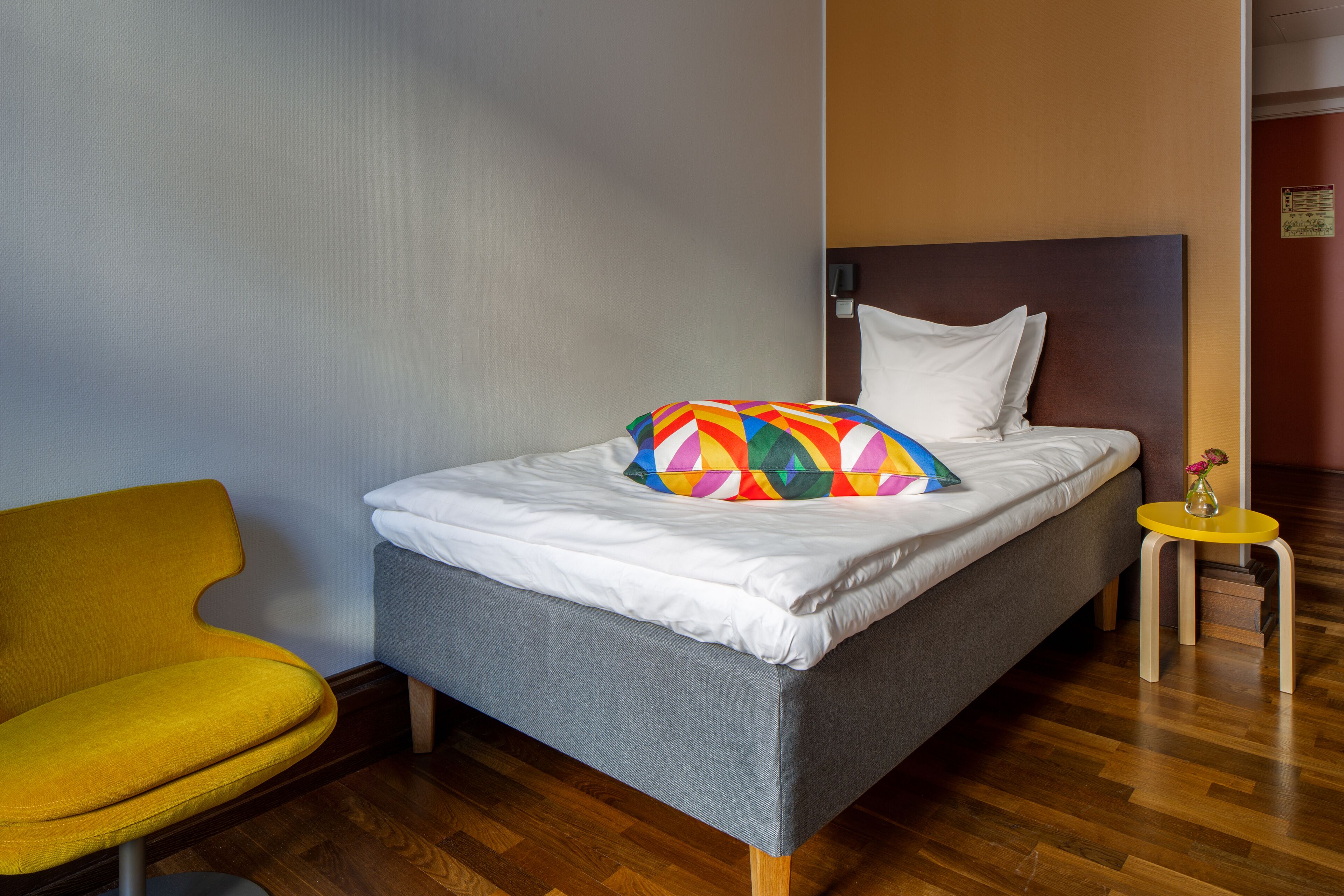 Una cama bien tendida con un cojín colorido, una mesita de noche de madera y una silla amarilla en una habitación de hotel moderna con suelo de madera.