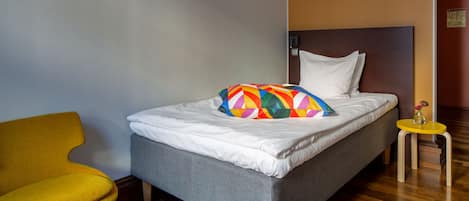 Una cama bien tendida con un cojín colorido, una mesita de noche de madera y una silla amarilla en una habitación de hotel moderna con suelo de madera.