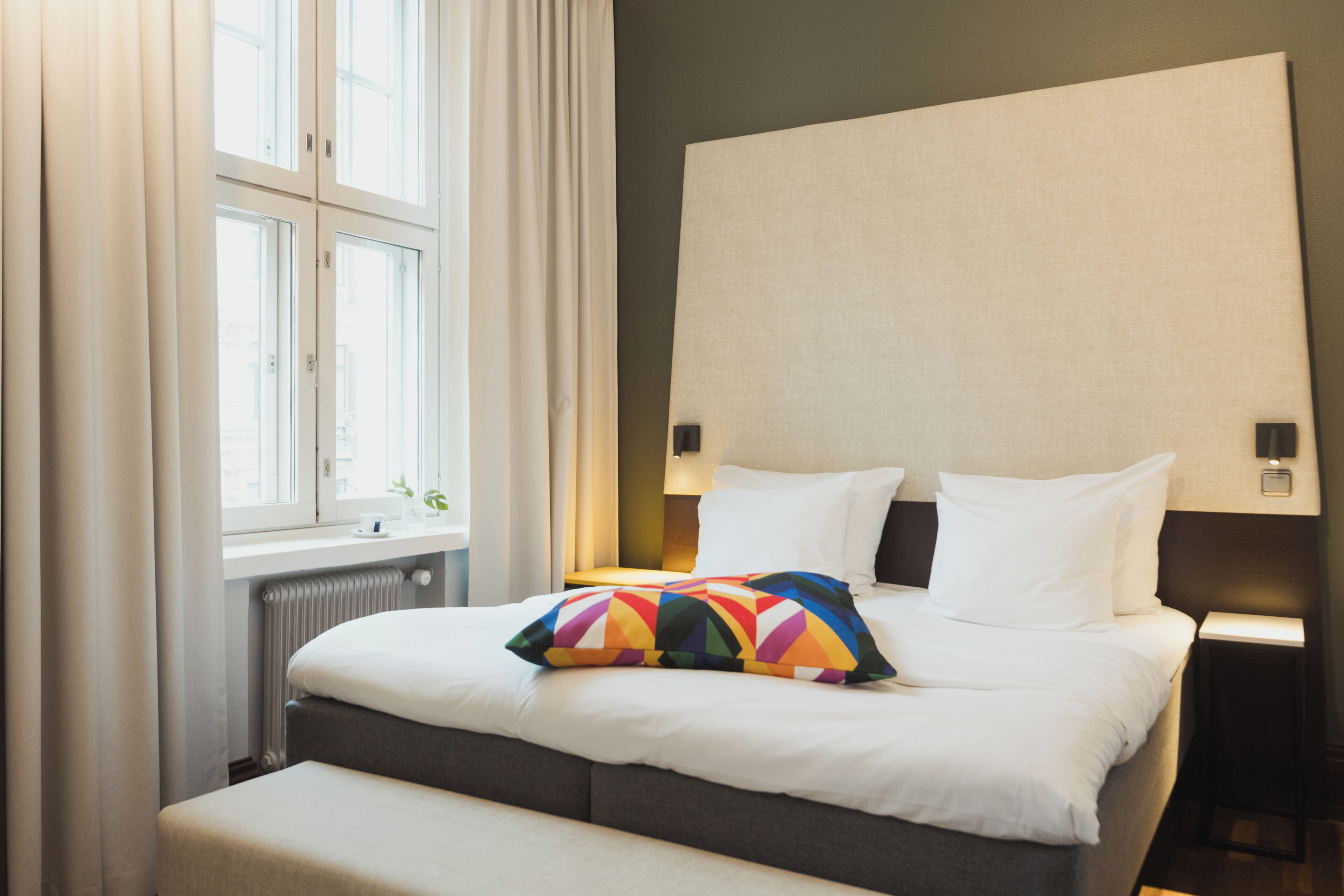 Un lit bien fait, avec du linge de lit blanc et un coussin aux motifs géométriques colorés, une lampe de chevet et une fenêtre avec des rideaux.
