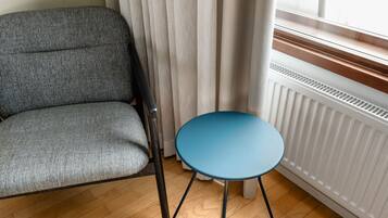 Een kamer met een grijs gestoffeerde fauteuil, een klein blauw tafeltje, een radiator en een raam met gordijnen.