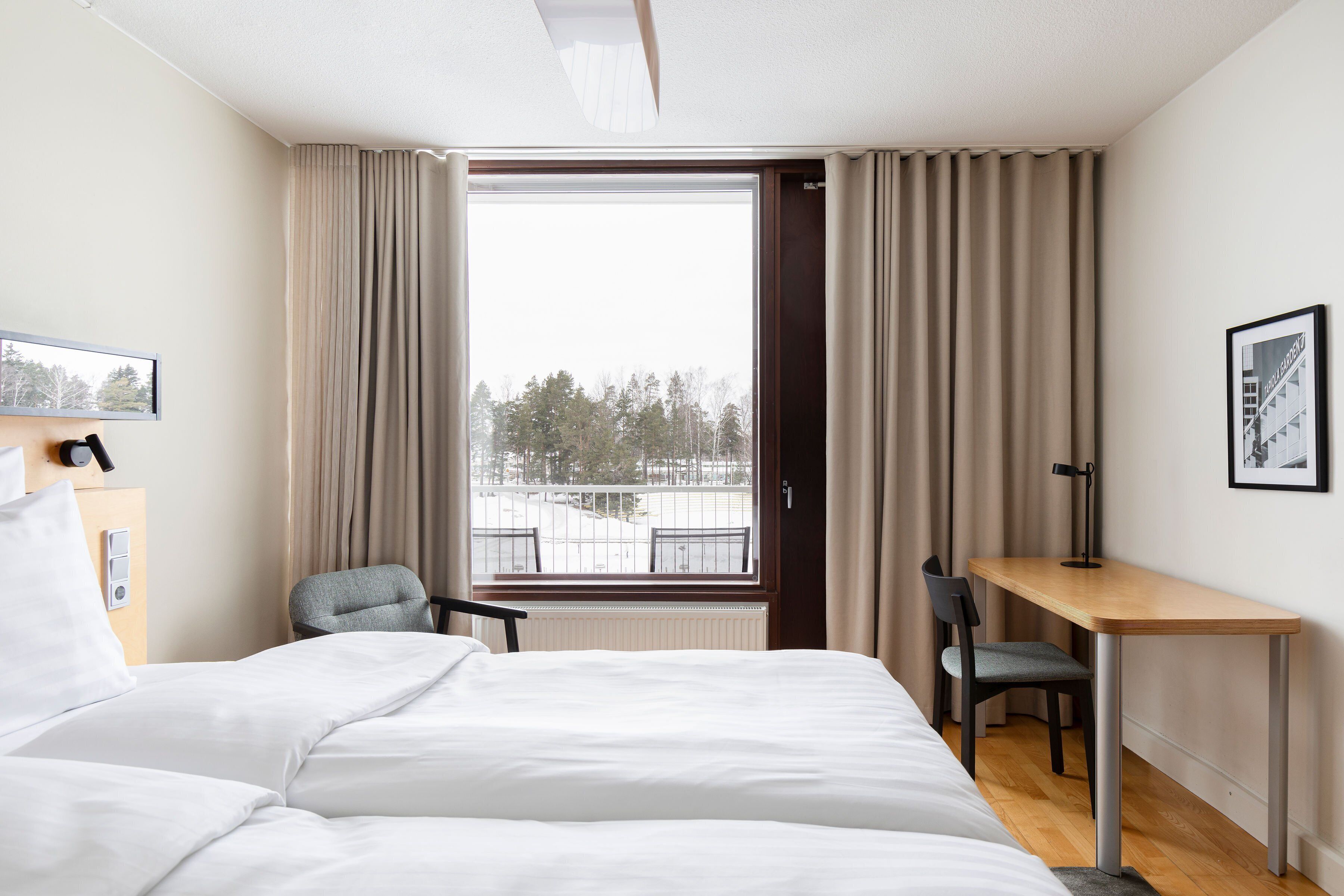 Habitación de hotel con cama, escritorio y silla. Se ve una ventana con cortinas, un radiador y un cuadro en la pared.