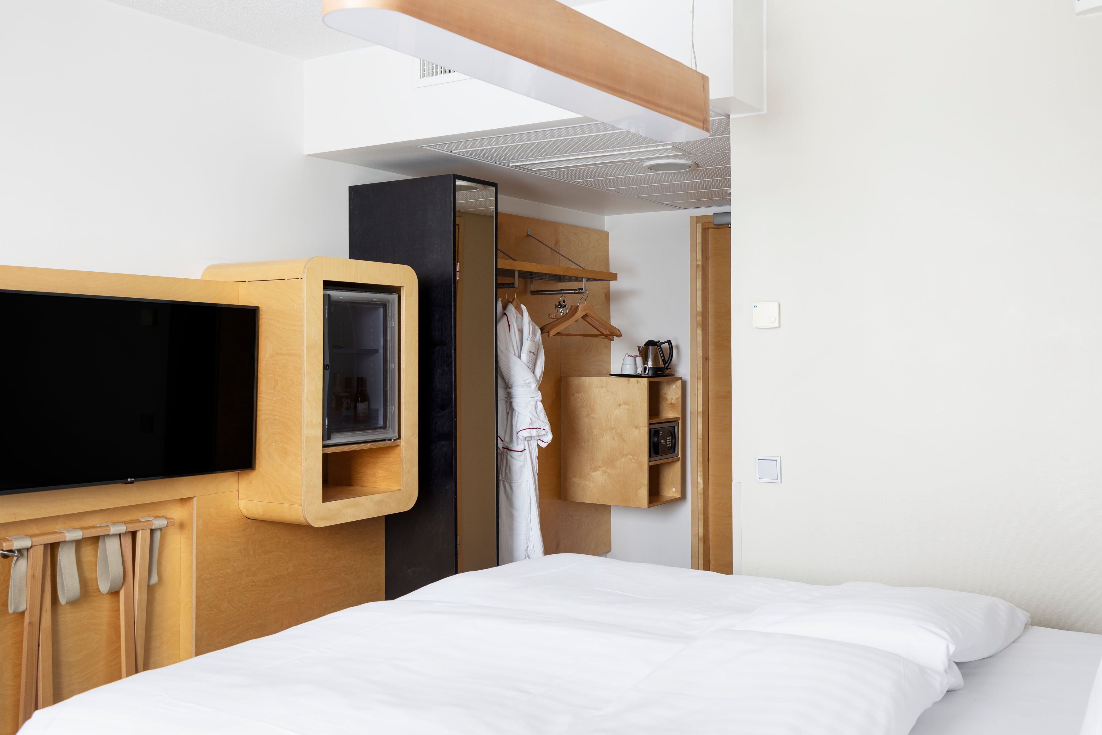 A modern hotel room with a large bed, a TV mounted on the wall, a wooden wardrobe, and a small shelf with toiletries.
