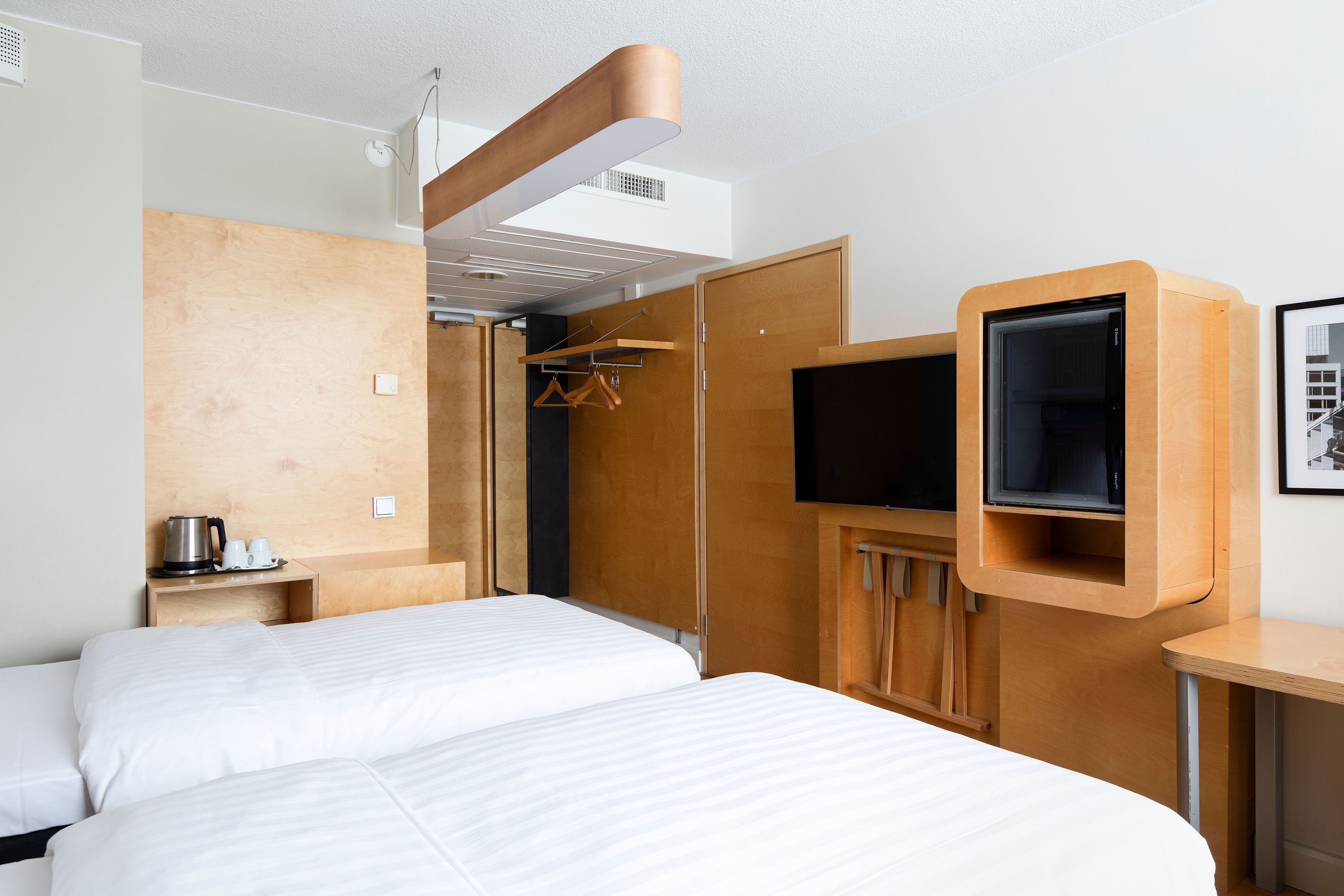 A hotel room with two beds, a wooden wardrobe, a TV mounted on the wall, and a small table with a kettle and cups.