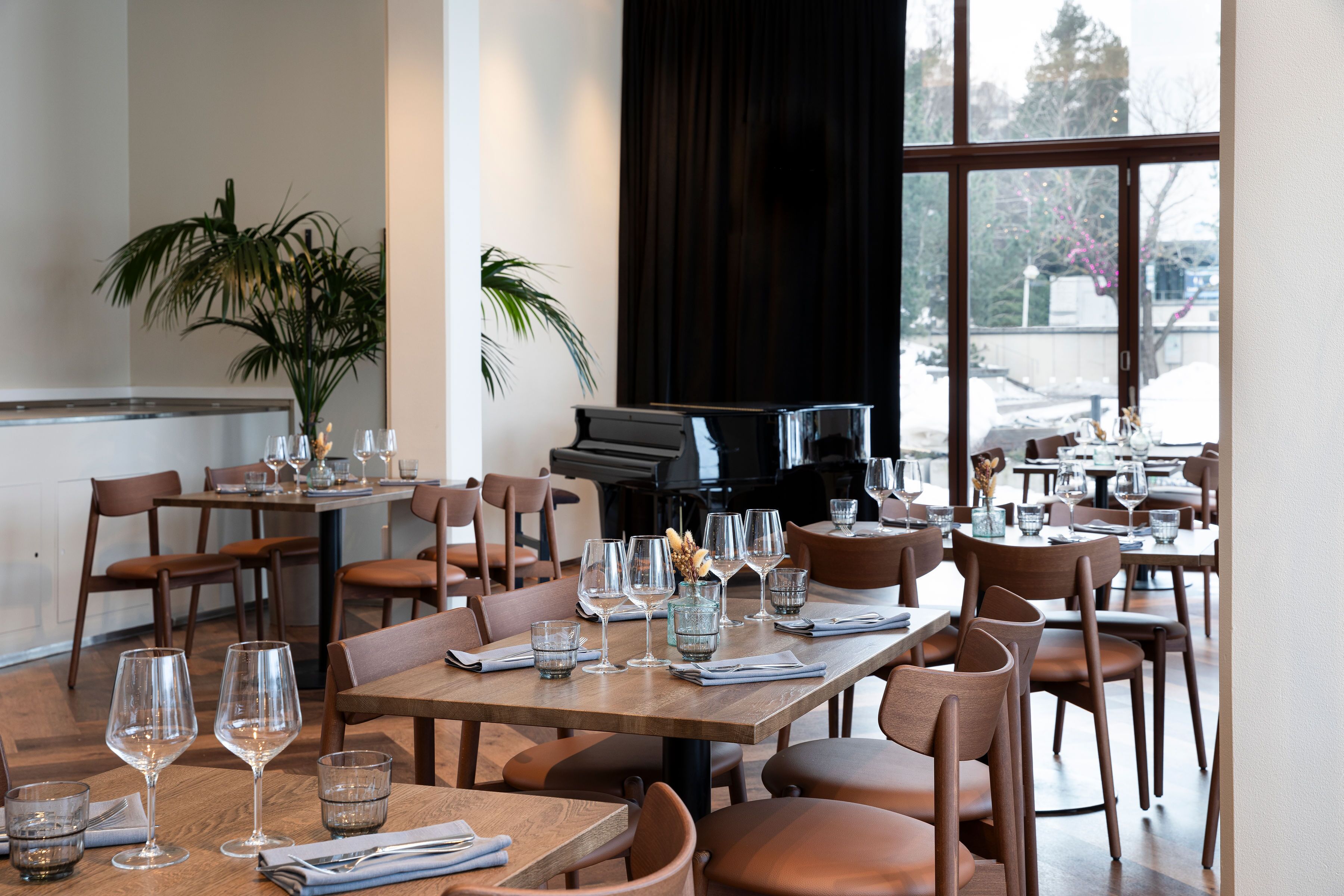 A restaurant with tables set for dining, a piano, and potted plants.