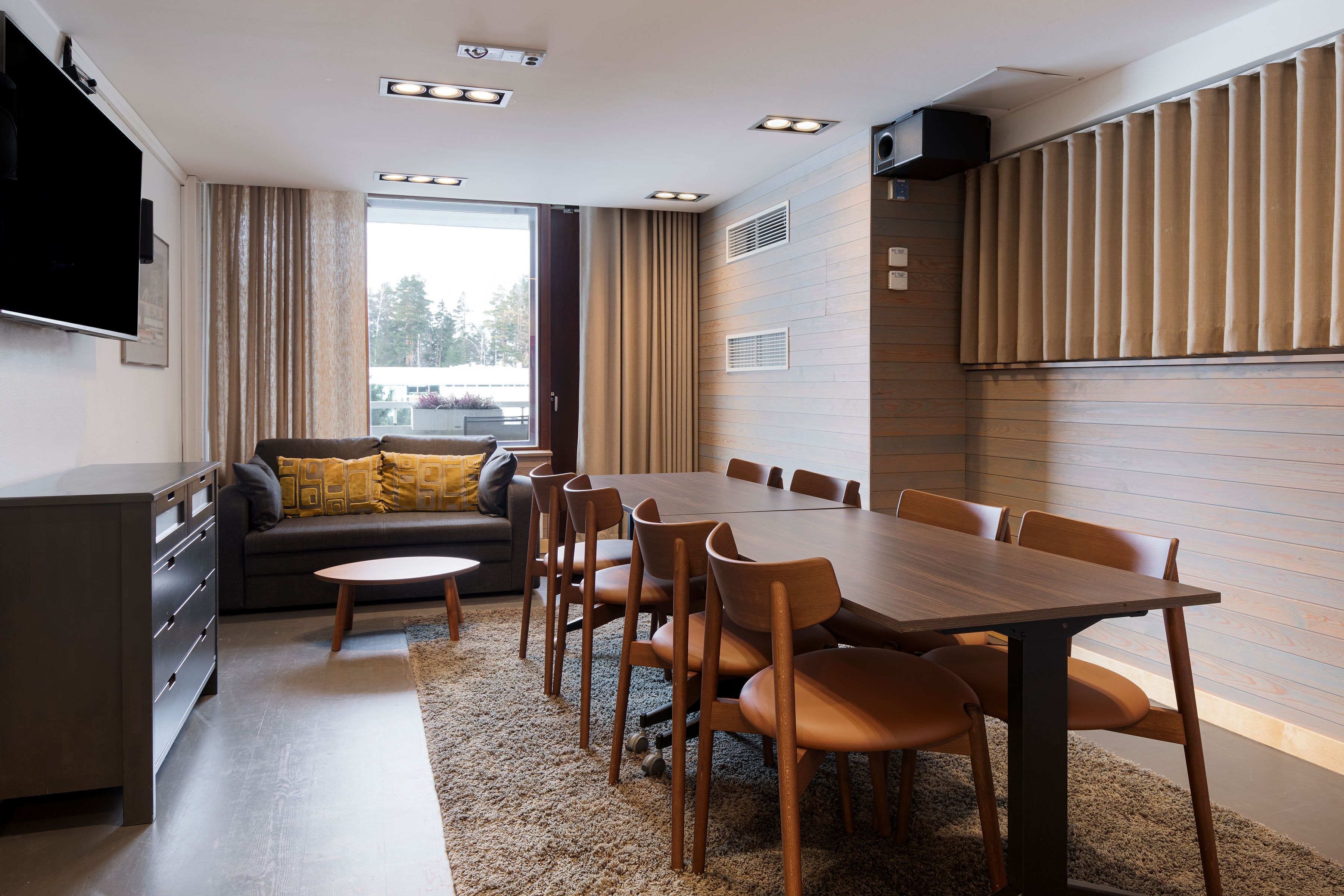 A modern dining area with a wooden table and chairs, a sofa with cushions, a flat-screen TV mounted on the wall, and a large window with curtains.