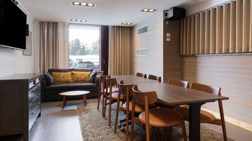 A modern dining area with a wooden table and chairs, a sofa with cushions, a flat-screen TV mounted on the wall, and a large window with curtains.