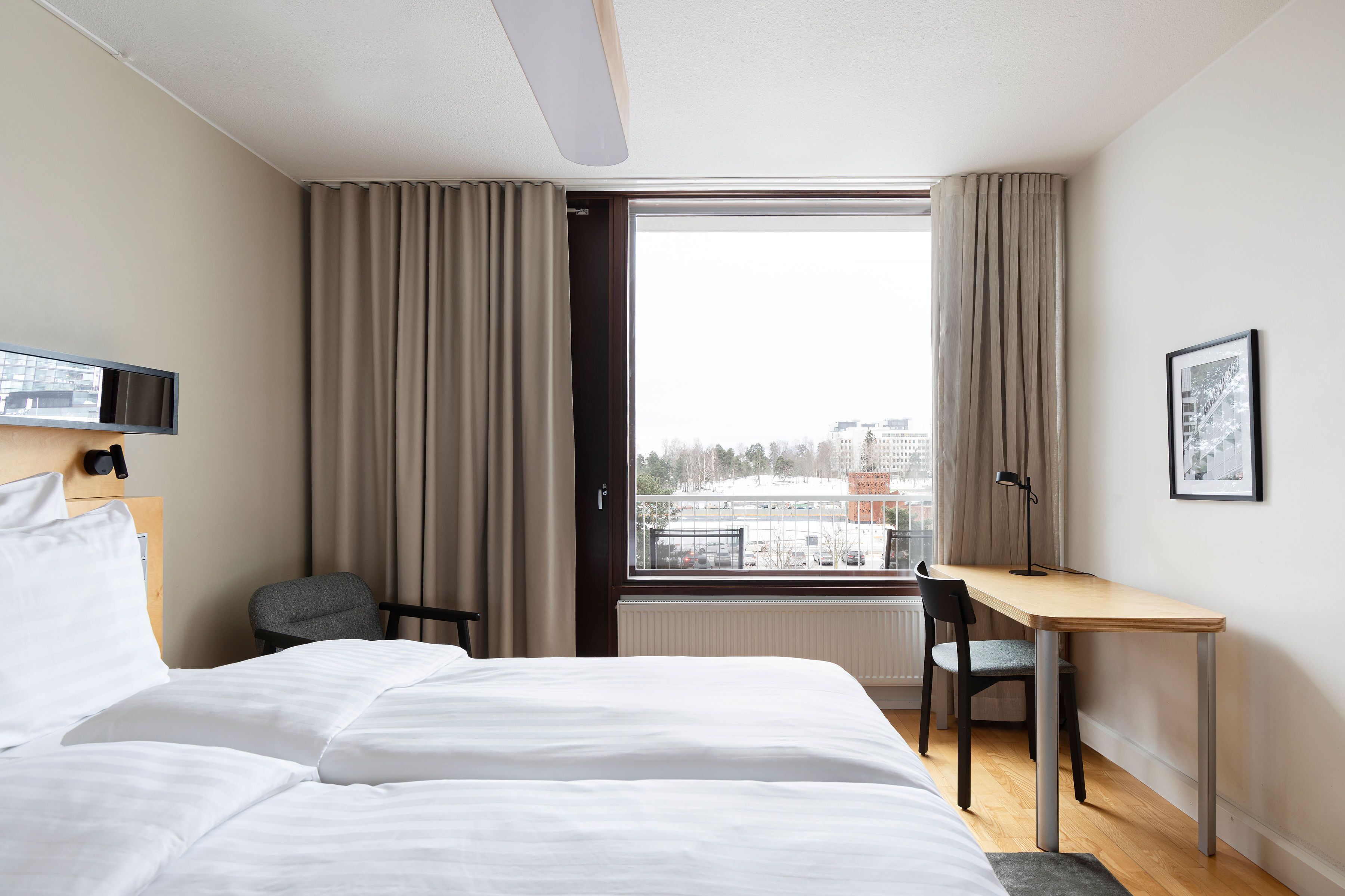 A hotel room with a bed, a desk, and a chair. There is a window with curtains, a radiator, and a picture on the wall.