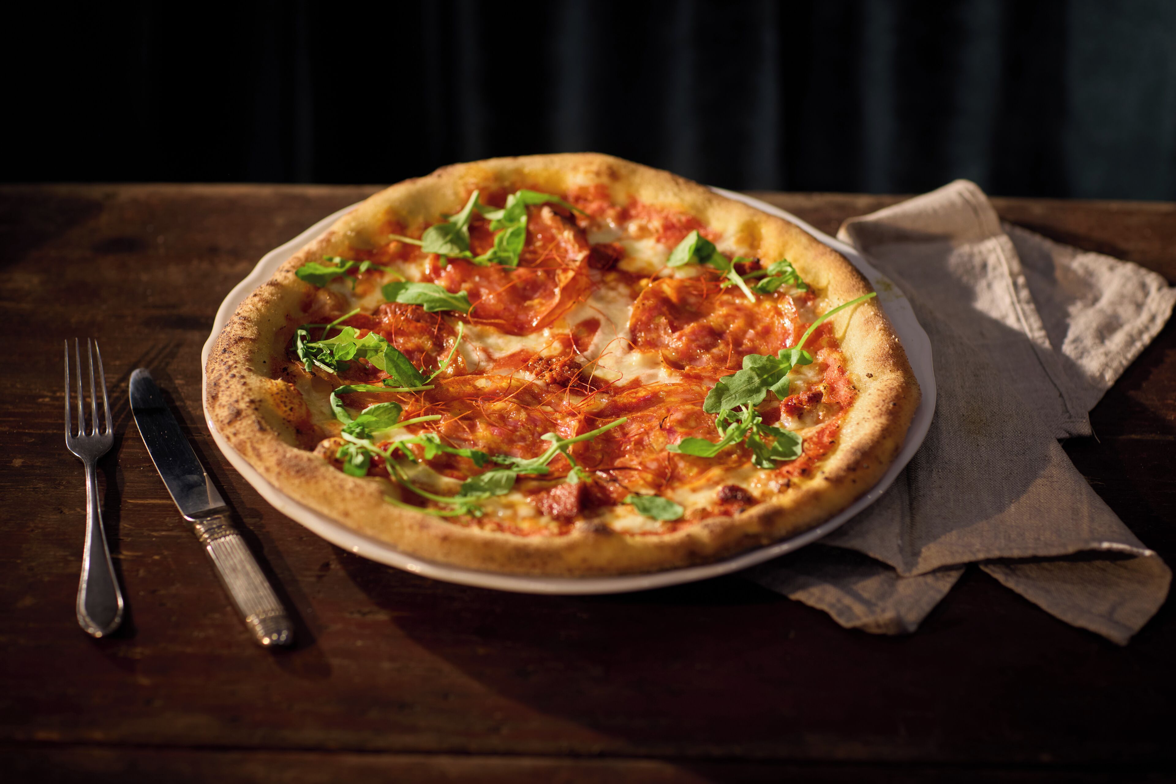 A pizza on a plate with a fork and knife beside it, placed on a wooden table with a napkin.