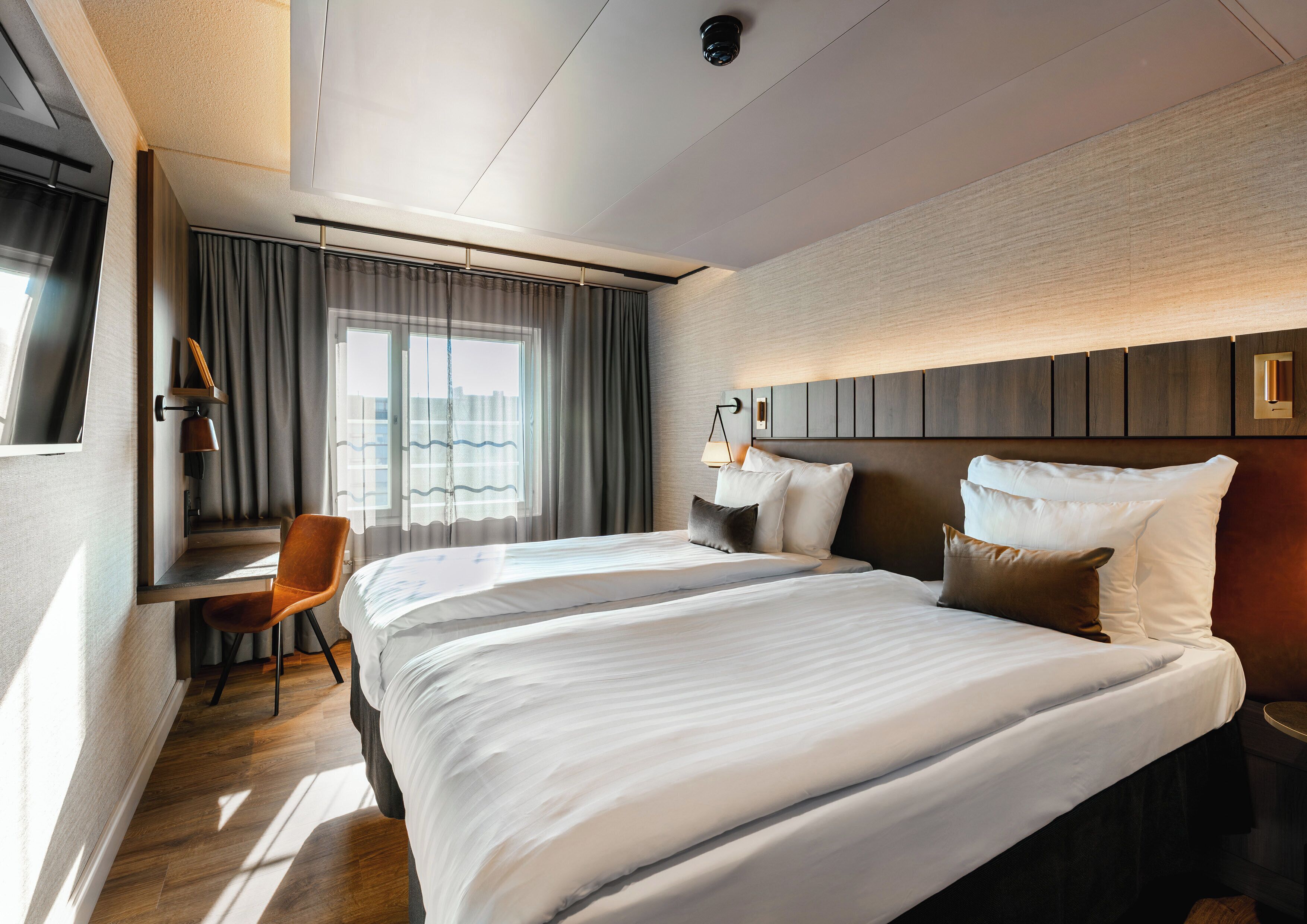 A hotel room with a large bed, a desk with a chair, a window with curtains, and a wall-mounted lamp.