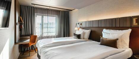A hotel room with a large bed, a desk with a chair, a window with curtains, and a wall-mounted lamp.