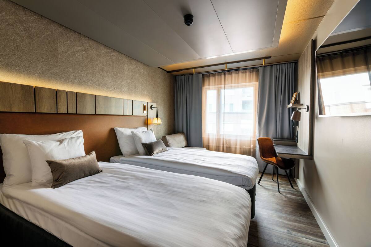 Ein Hotelzimmer mit zwei Betten, einem Schreibtisch, einem Stuhl, einem großen Fenster mit Vorhängen und einer Wandlampe.