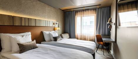 A hotel room with two beds, a desk, a chair, a large window with curtains, and a wall-mounted lamp.