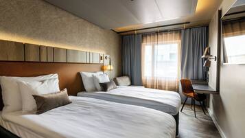 A hotel room with two beds, a desk, a chair, a lamp, and a view of the city through large windows.