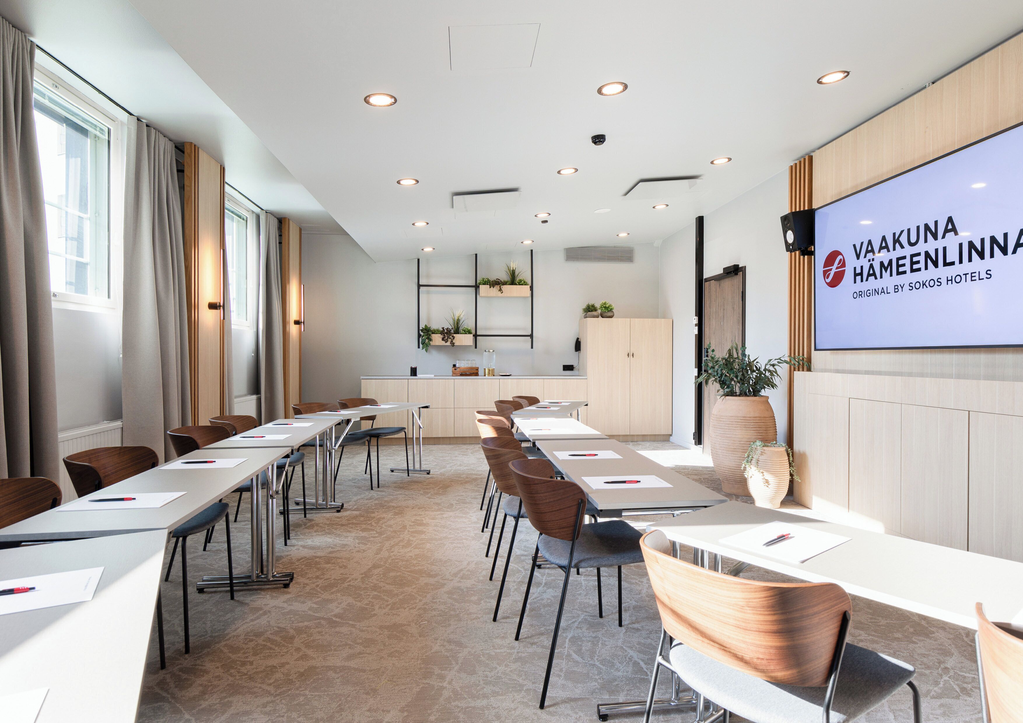 A modern conference room with long tables, chairs, and a large screen displaying the hotel's name.