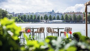 Une terrasse au bord de la rivière, agrémentée de chaises en osier et de plantes en pot, d’où l’on peut admirer un panorama sur la ville, avec ses bâtiments et le fleuve.